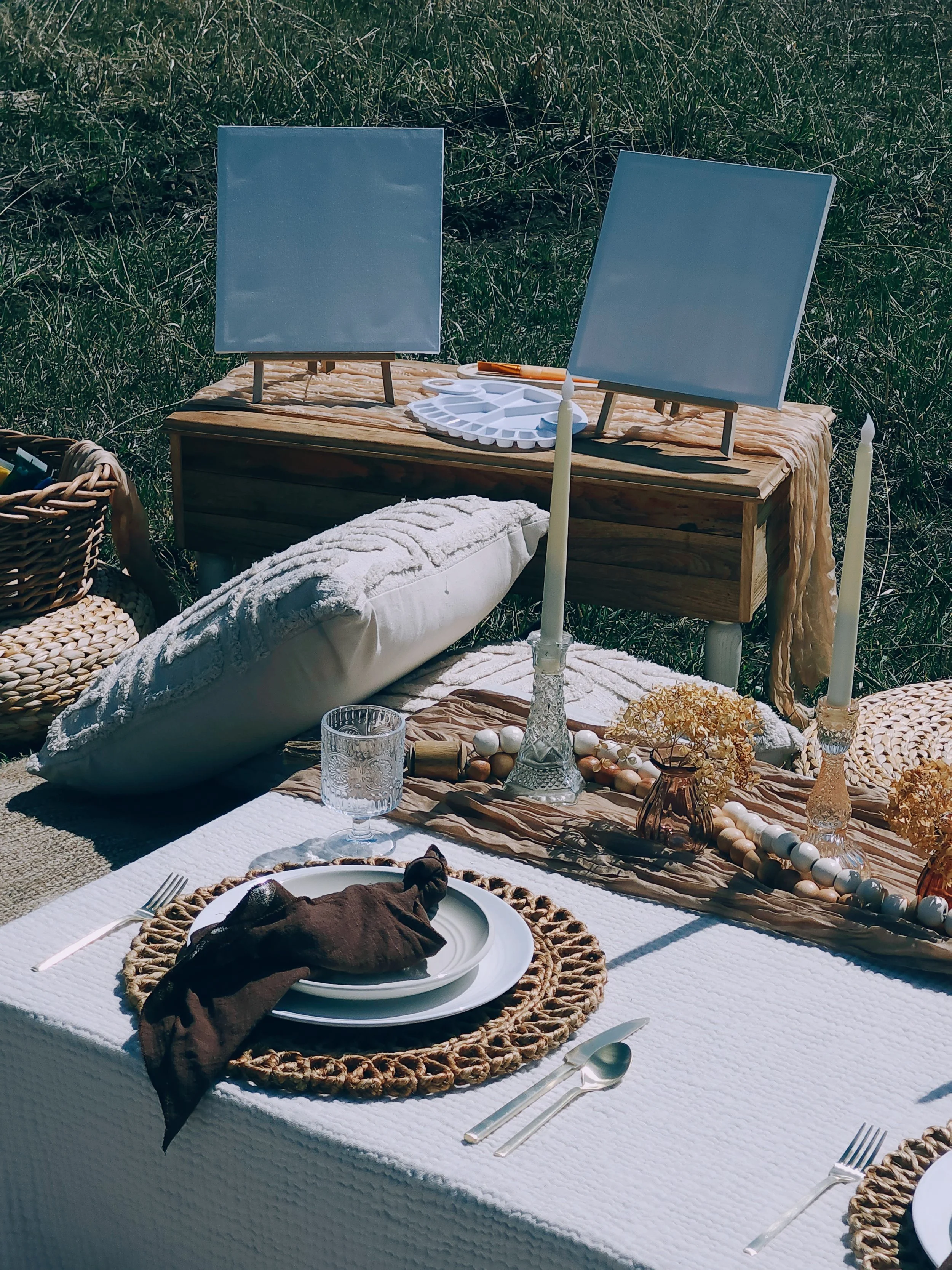 Outdoor table set for a meal with white plates, brown napkins, glassware, candles, and decorative beads, with paintings on easels in the background and natural greenery.