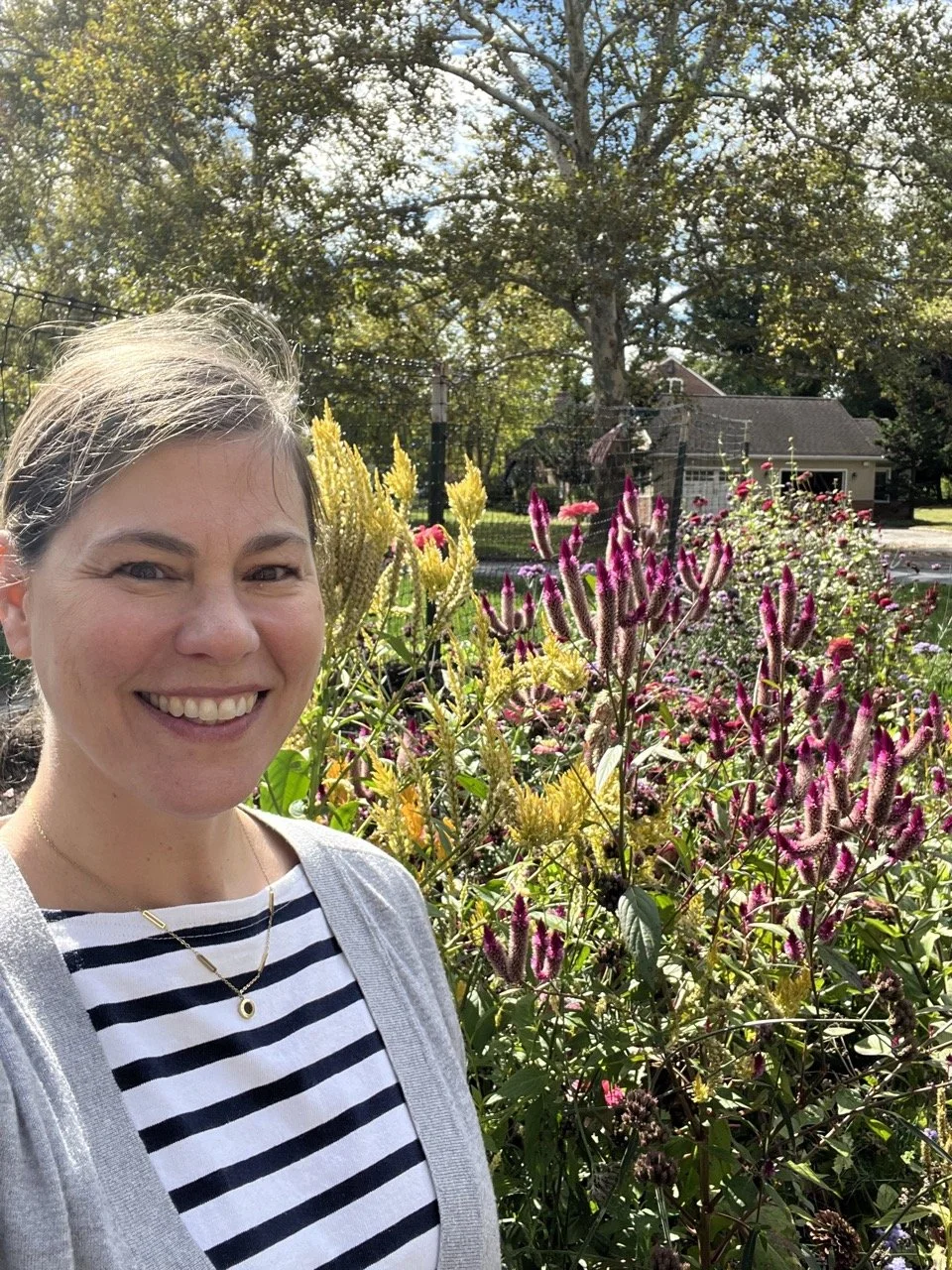 A smiling woman outdoors in a garden with flowers, trees, and a house in the background on a sunny day.