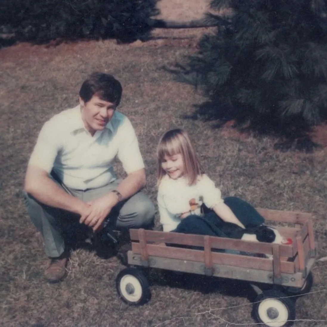 A man and a young girl behind a small wooden wagon with a black rabbit inside, outdoors on grass with bushes in the background.