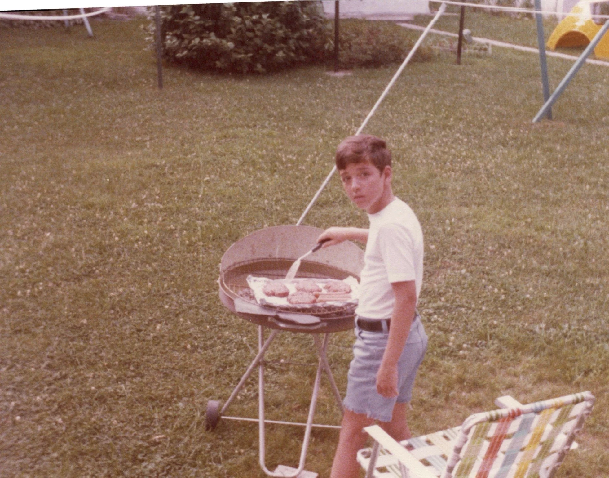 A young boy standing on a grassy yard, holding a folding table with food on it. There is a patio table with an umbrella, a lawn chair, and playground equipment in the background.