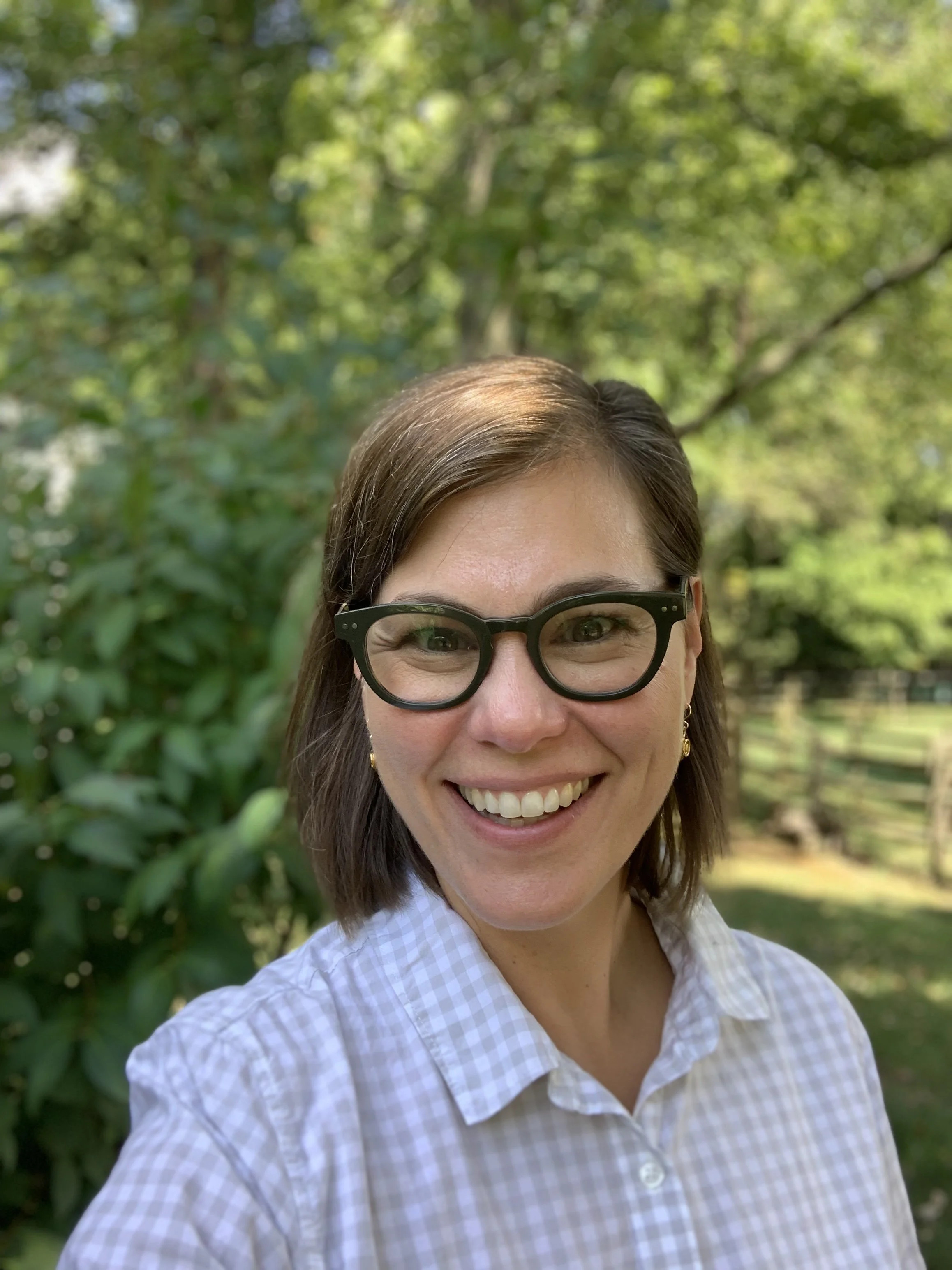 A woman with brown hair, glasses, and earrings smiling outdoors with green trees and foliage in the background.