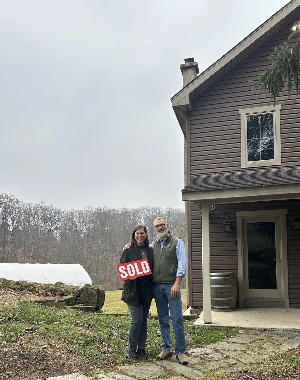 Two people standing outside a house, smiling, holding a red 'SOLD' sign, with a backyard and a greenhouse in the background.