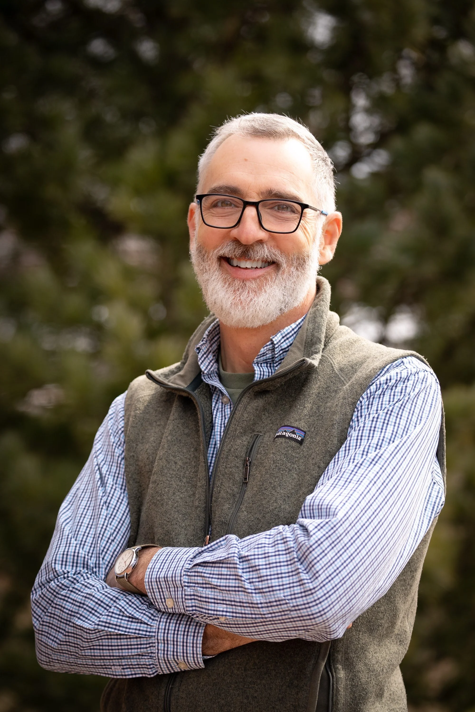A middle-aged man with glasses and a beard, wearing a plaid shirt and an outdoor vest, smiling while outdoors.