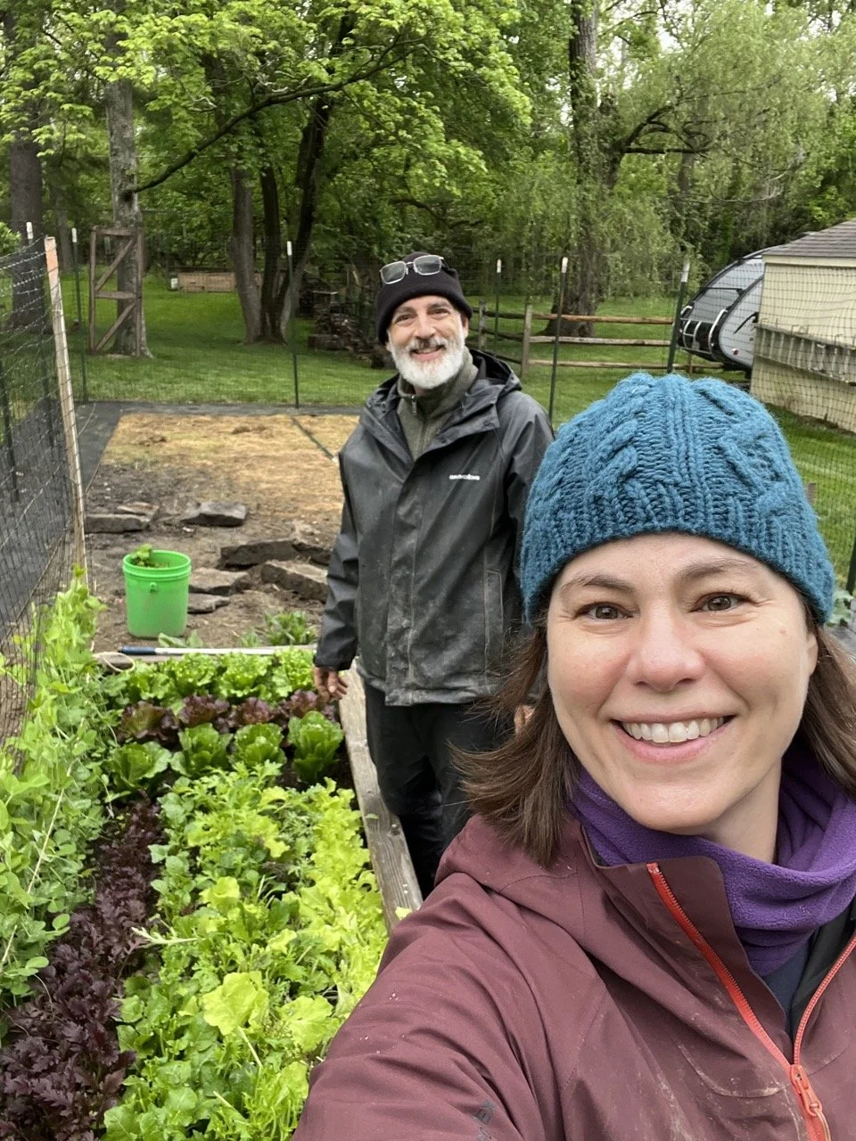 A smiling woman in a purple jacket and blue knit hat taking a selfie in a vegetable garden, with a smiling man in a black jacket and beanie standing behind her, surrounded by green trees and outdoor fencing.