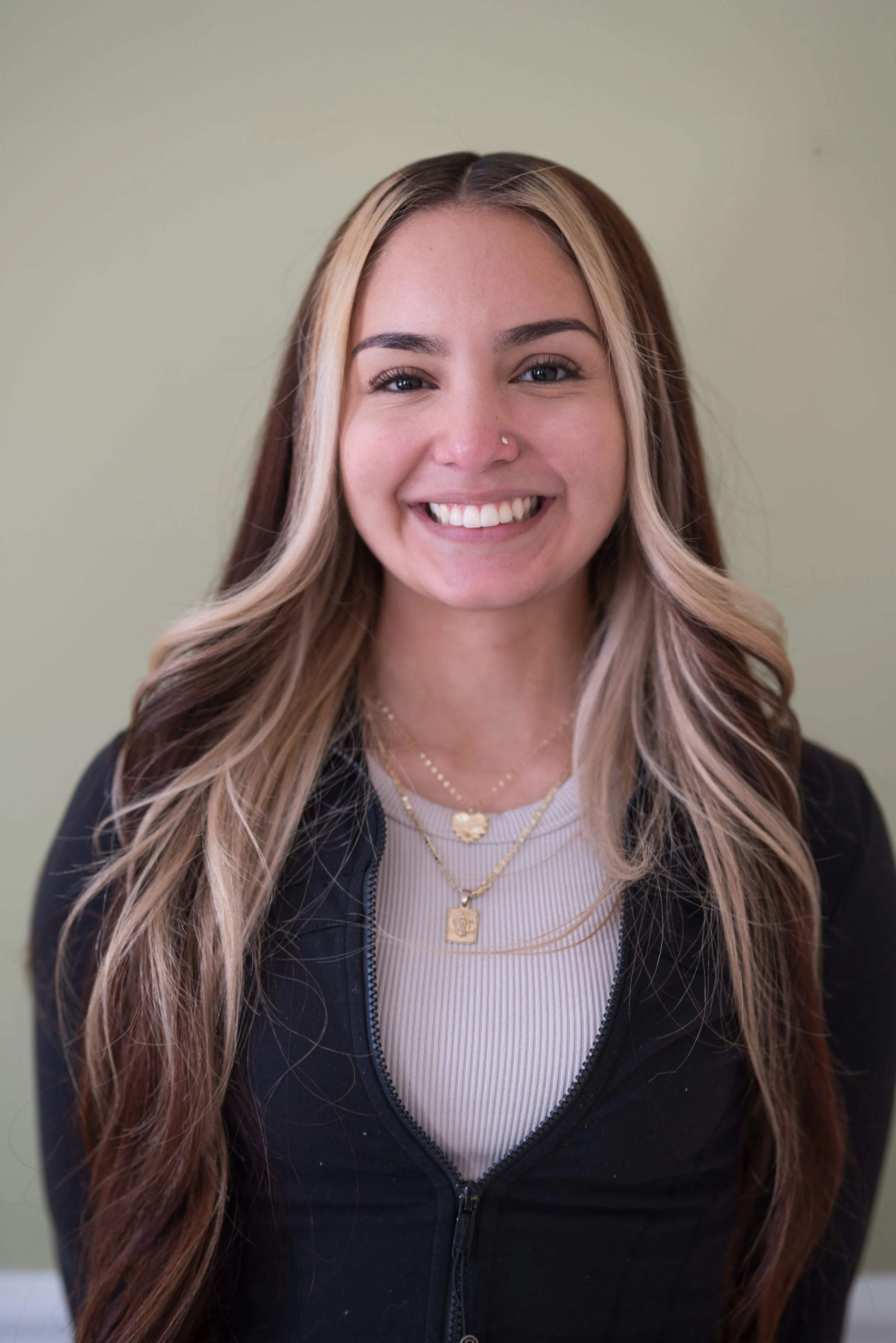 A smiling young woman with long, wavy hair that has blonde highlights, wearing layered necklaces, a light-colored ribbed top, and a black zip-up jacket, standing against a plain light green wall.