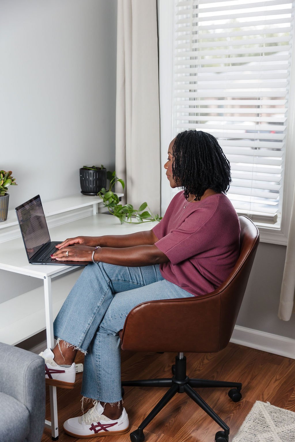 A woman with black hair, pink shirt, blue jeans, and sneakers typing on a laptop