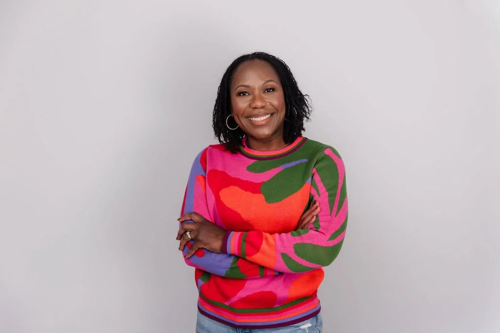 A woman with black hair, smiling, wearing a colorful sweater with pink, red, green, purple, and orange with a plain light gray background.