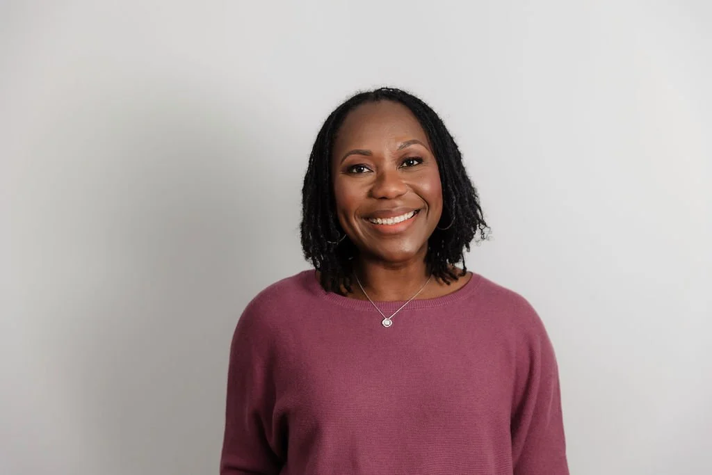 A woman with black hair, pink shirt, and a necklace smiling with grey background