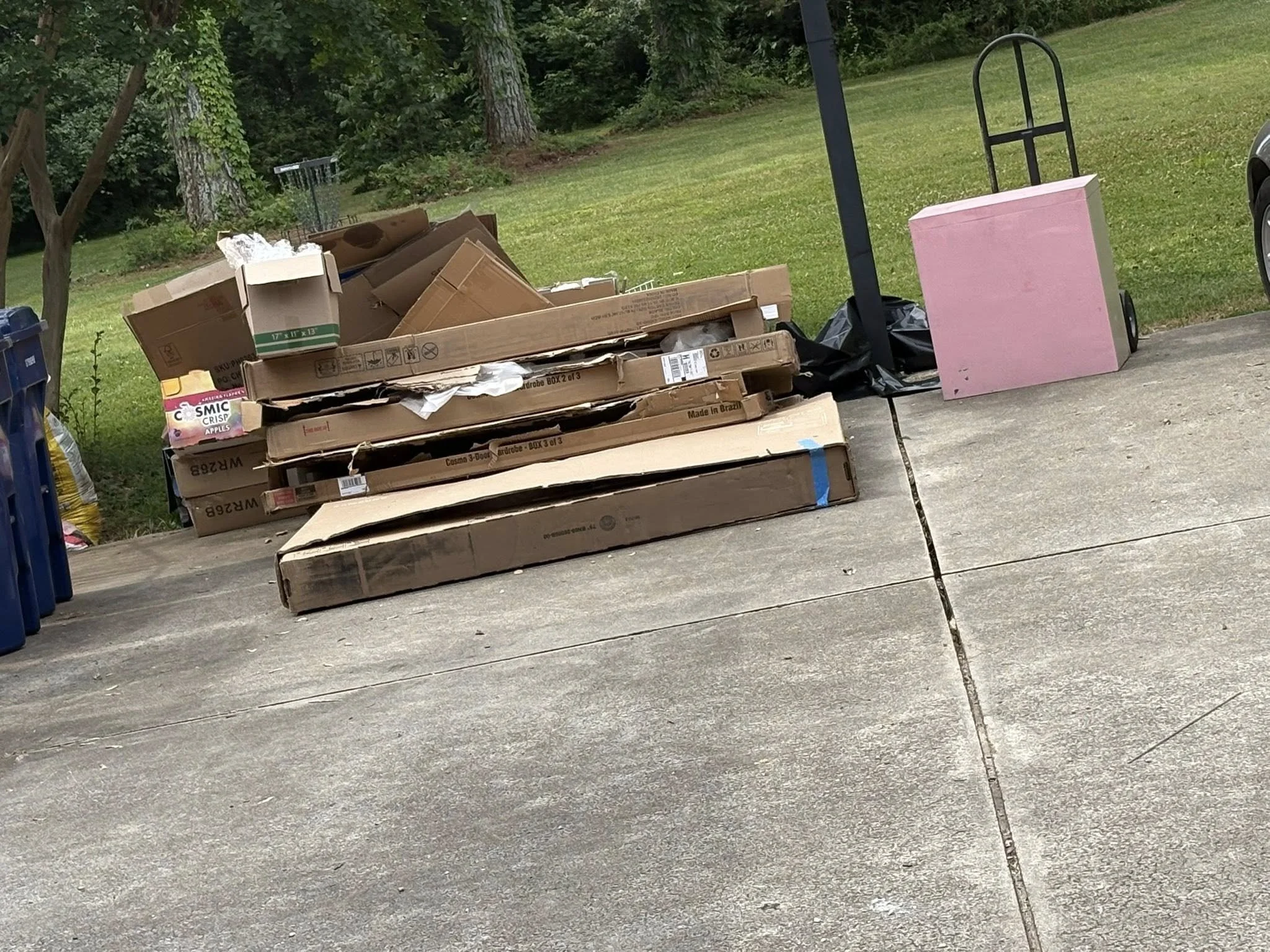 A pile of cardboard boxes on a concrete driveway, with some boxes flattened and others stacked on top of each other. To the right, there is a pink foam board leaning against a black hand truck. The background features green grass, trees, and bushes.