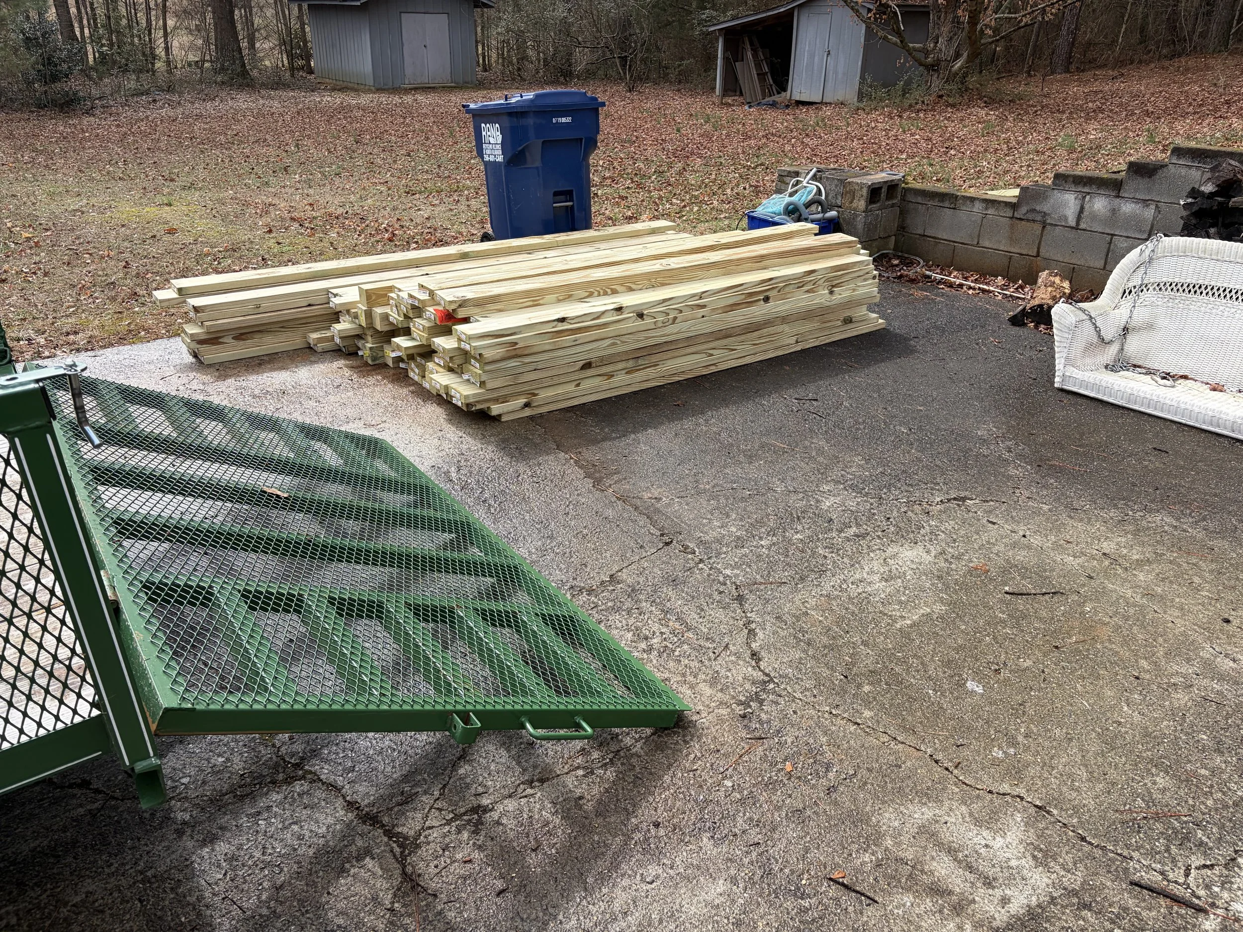 Stacked lumber, a blue garbage bin, a green metal cart, a white wicker bench, and outdoor items on a concrete driveway with a wooded background.