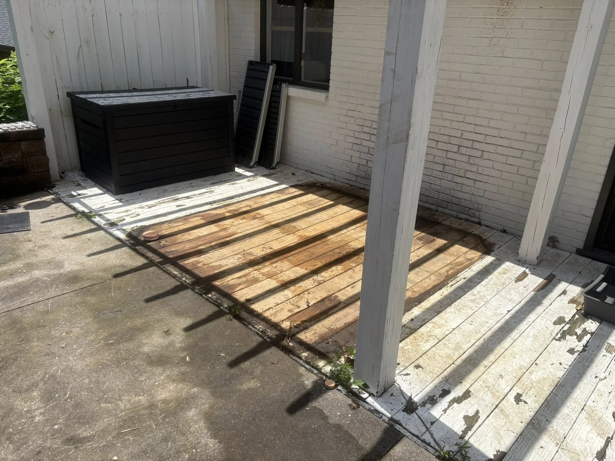 A small backyard patio area with white brick walls and a white wooden fence. There is a black storage box and two folding chairs leaning against the wall. The deck is partially covered with a wooden surface, and the area is partially shaded with sunl