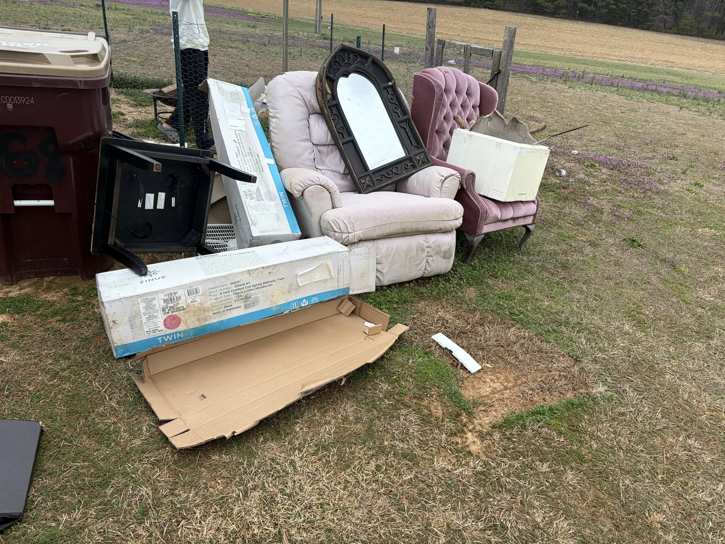 Discarded furniture and household items on a patch of grass outdoors, including two armchairs, a mirror, a small black table, a large white box, and flattened cardboard boxes, with a barn and fence in the background.