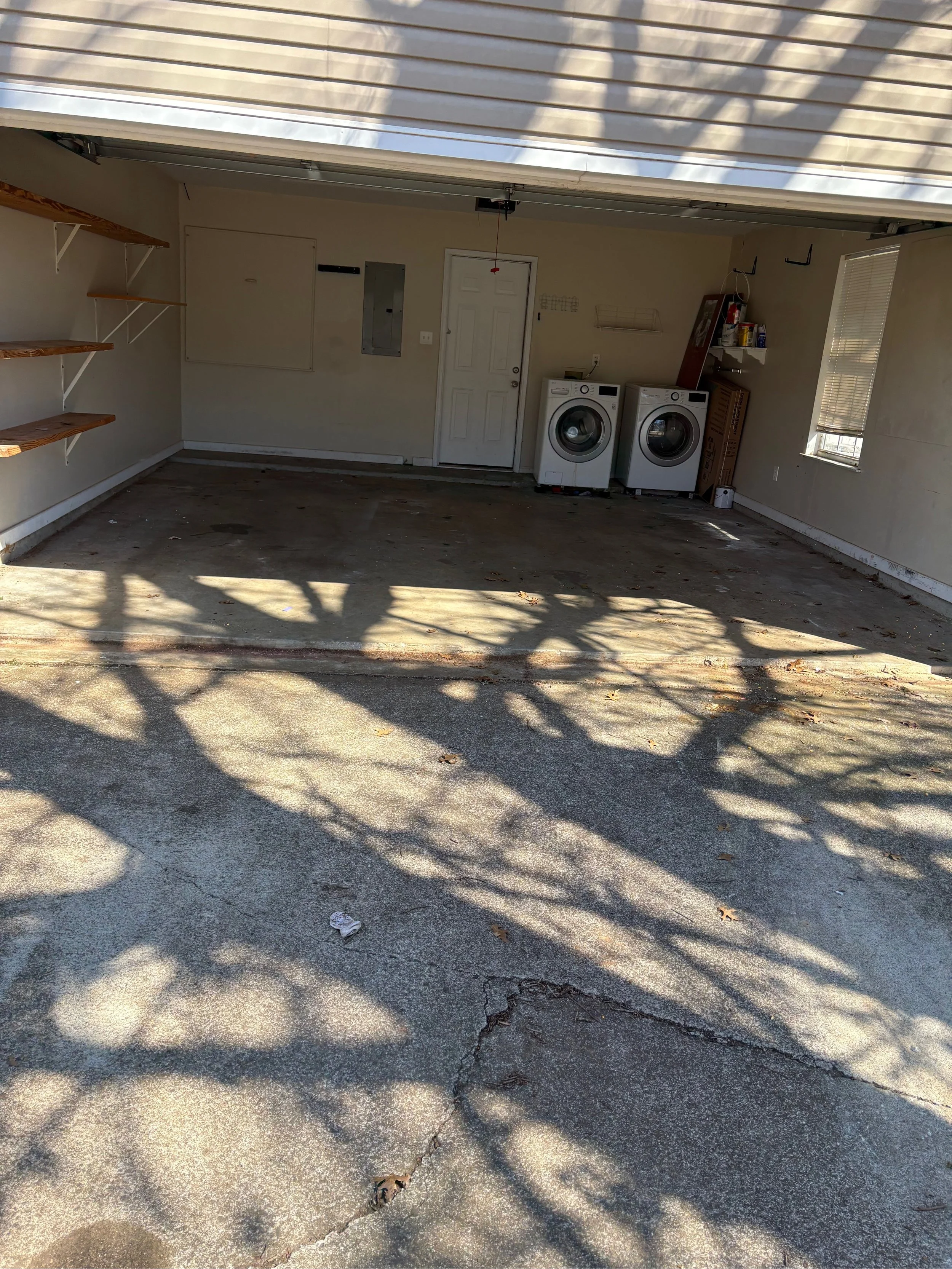 Empty garage with washer and dryer, wooden shelves on the left wall, and a window on the right wall, with sunlight and shadows on the concrete floor.