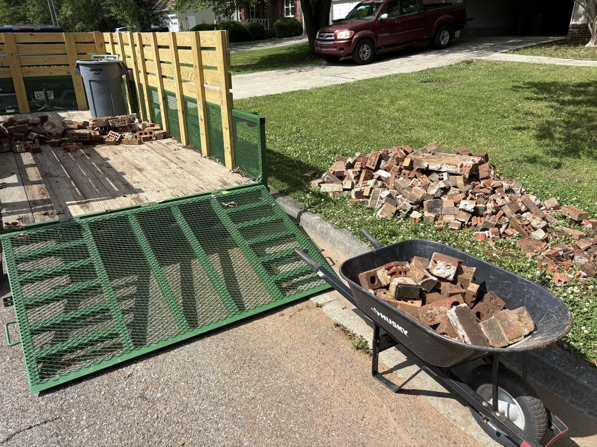 Pile of broken bricks in a wheelbarrow on a driveway next to a yard with a wooden fence under a tree. A red vehicle is parked in the driveway of a house in the background.