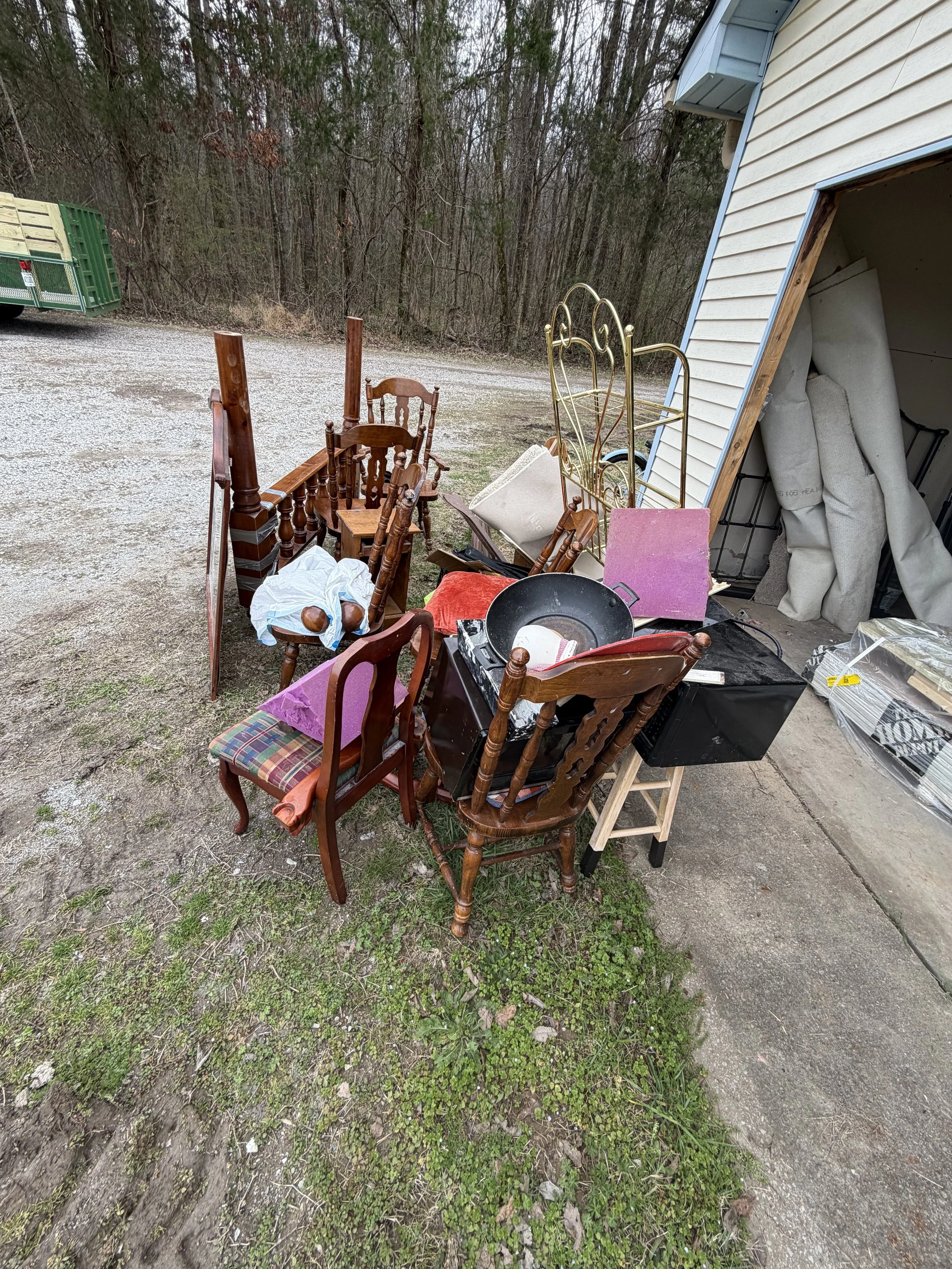Various pieces of furniture and household items, including wooden chairs, a metal headboard, a lamp, and boxes, are scattered outside a garage with some fabric or padding leaning against the wall surrounded by a wooded area.