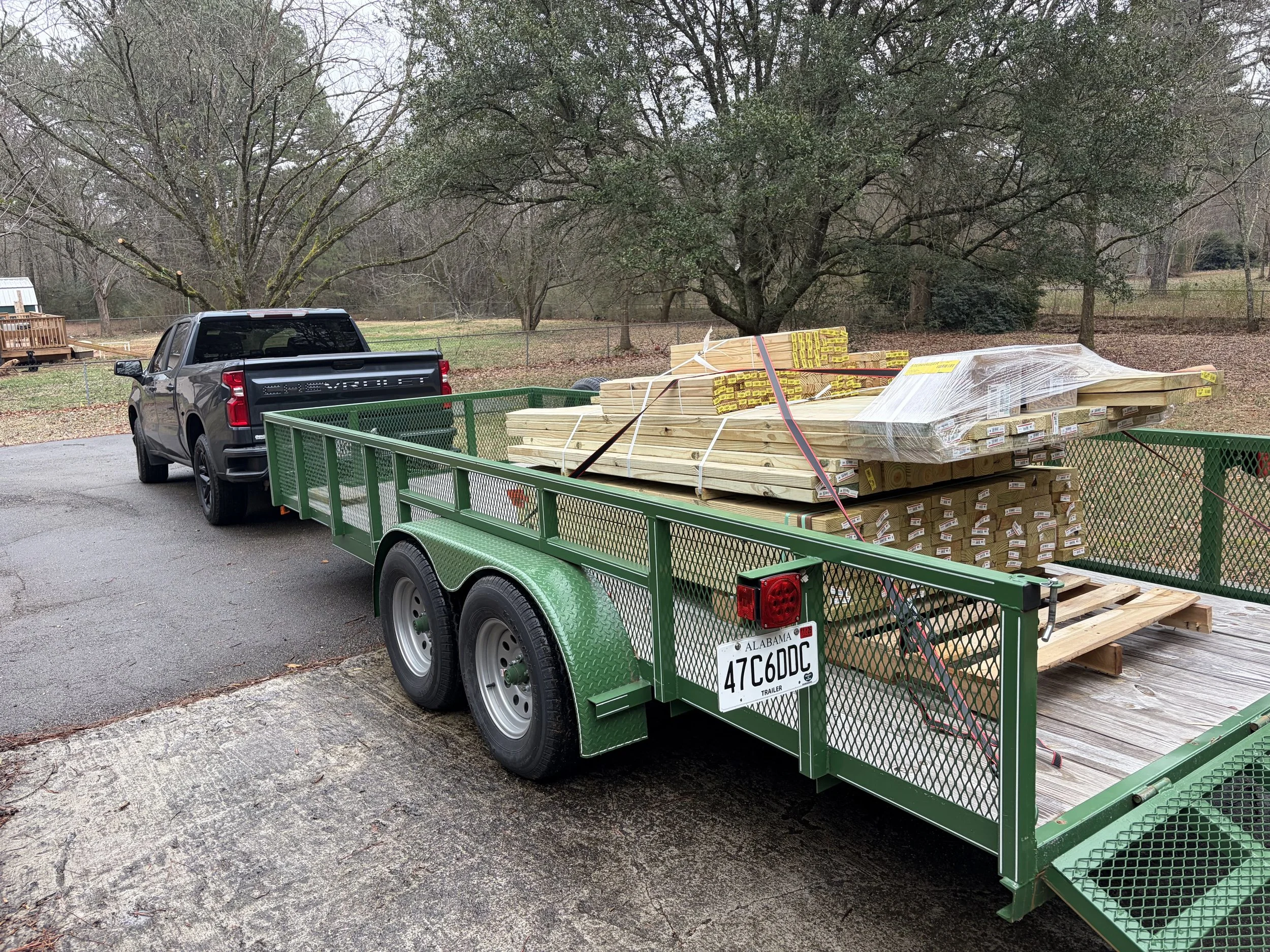 Green utility trailer attached to a black pickup truck, loaded with wood planks, wrapped with straps, parked on a paved driveway in a residential backyard with trees and grass.