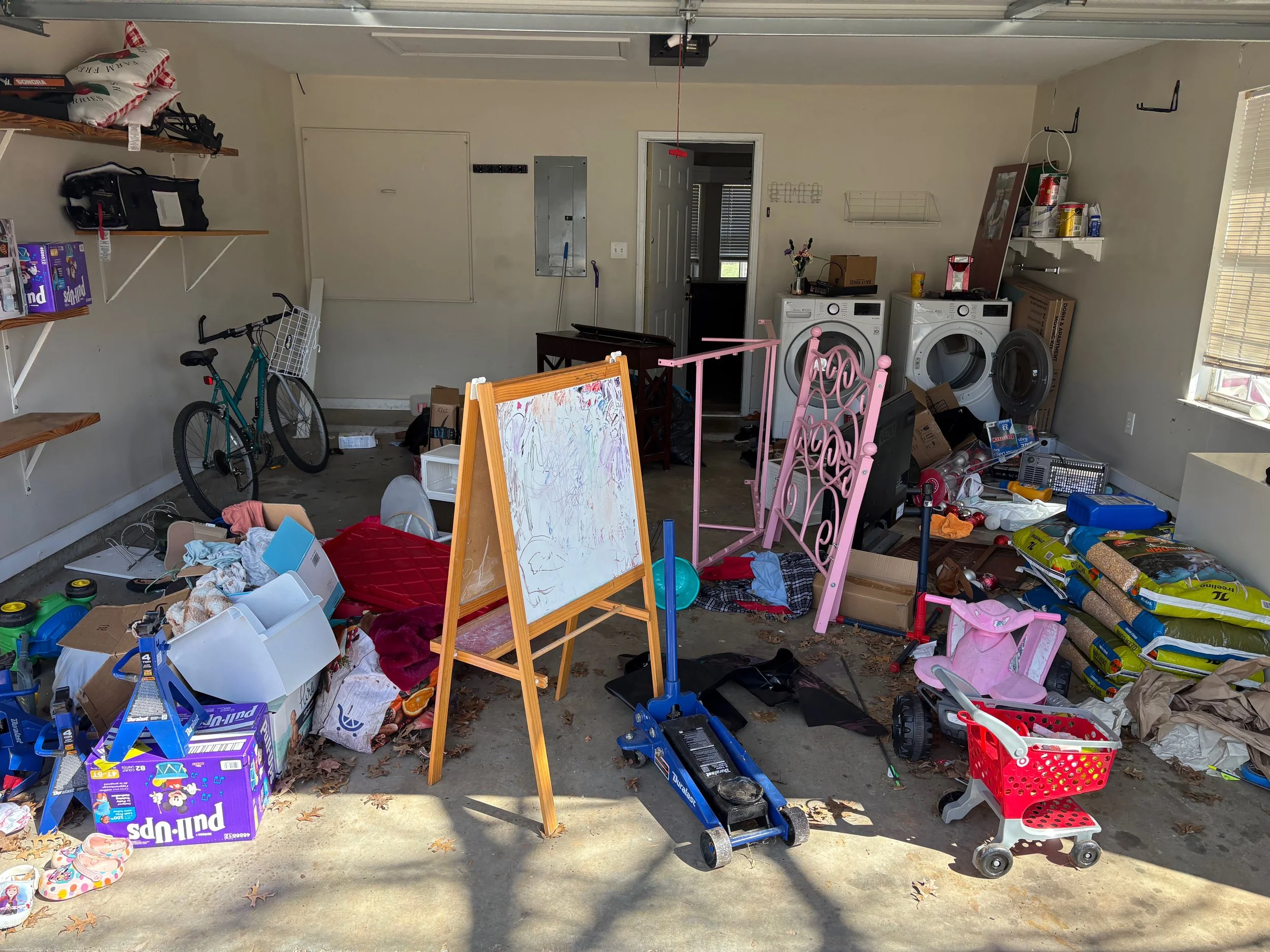 A cluttered garage with various toys, boxes, and household items scattered around, including a bicycle, a pink toy cart, a blue toy hand truck, washing machines, and a small whiteboard. There are shelves with miscellaneous items and a window with bli