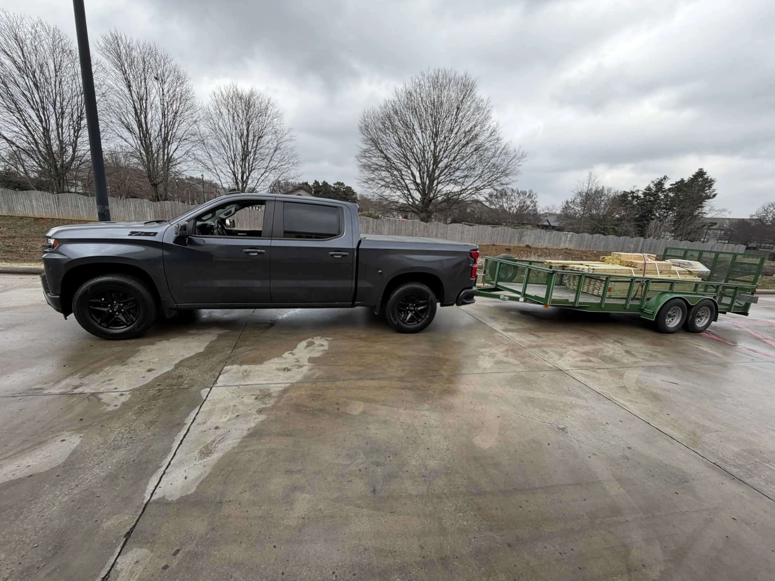 A black pickup truck pulling a green dual-axle trailer loaded with wooden pallets on a concrete surface with leafless trees and a cloudy sky in the background.