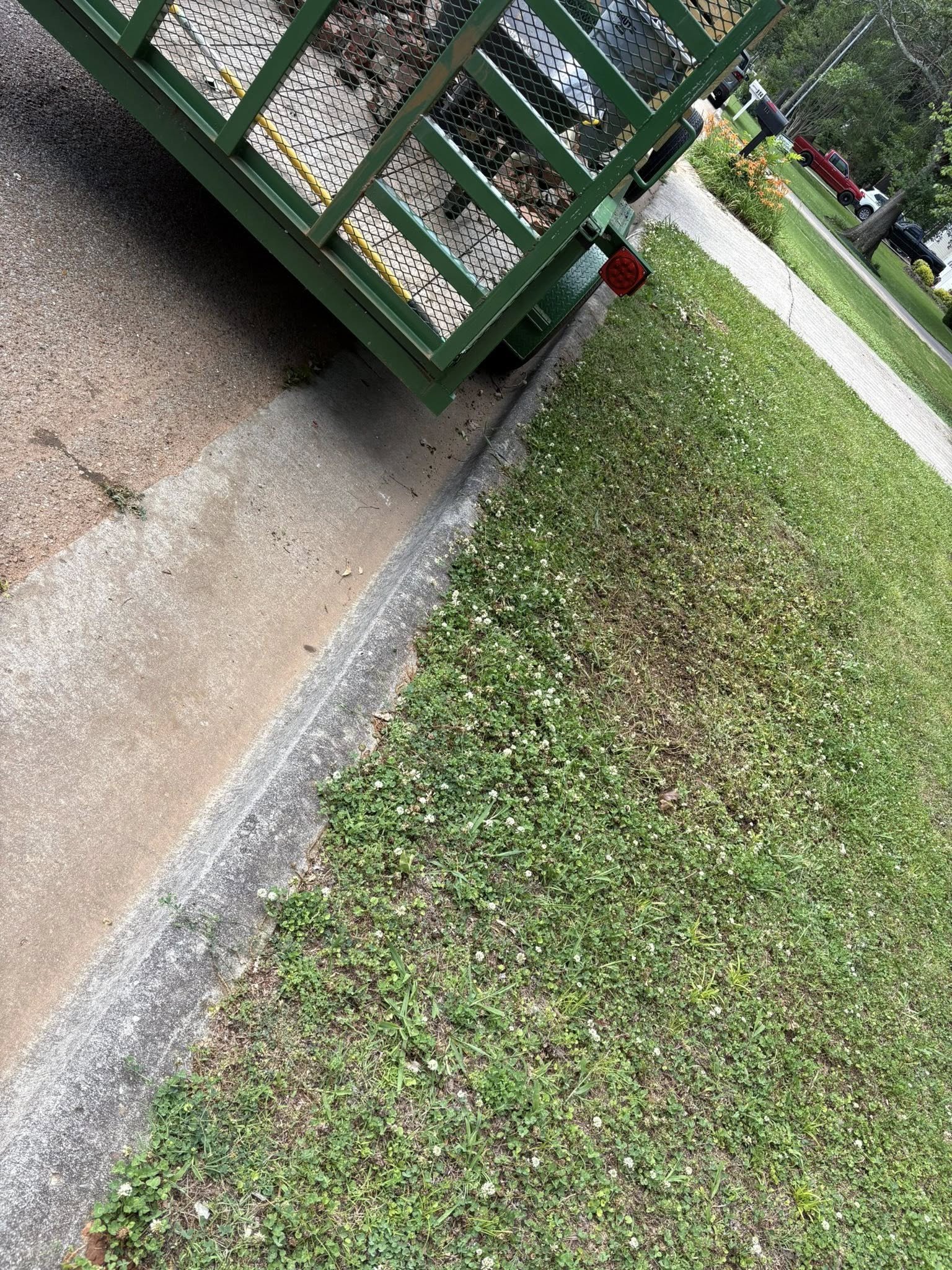 A green metal utility trailer parked on a sidewalk near a grassy area with small white flowers. The trailer contains stacked firewood and is positioned next to a curb with a gravel and dirt path.