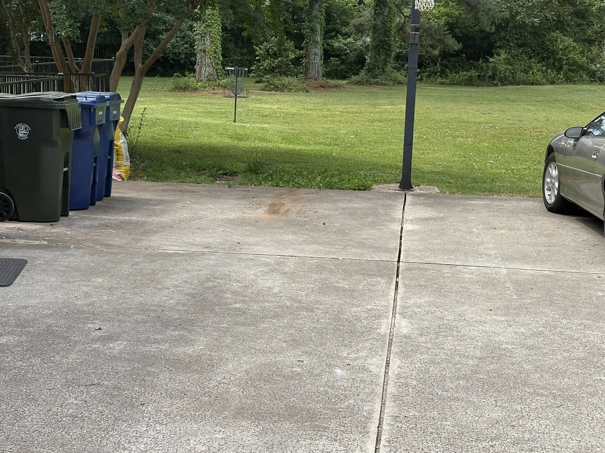 Backyard with green grass, trees, a basketball hoop, and several trash and recycling bins in the foreground.