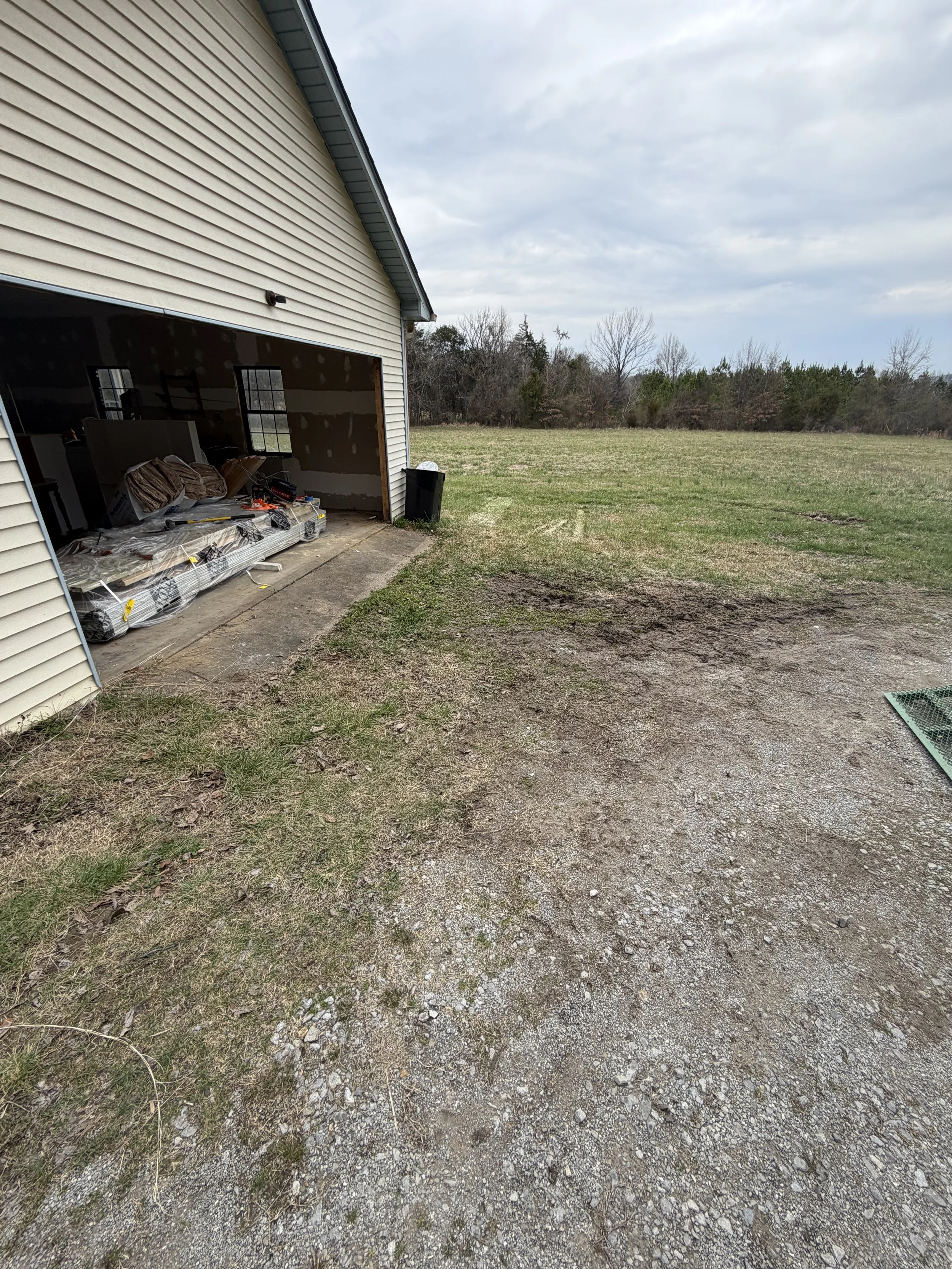 View of an open garage showing construction materials, with an open grassy yard and trees in the background under a cloudy sky.