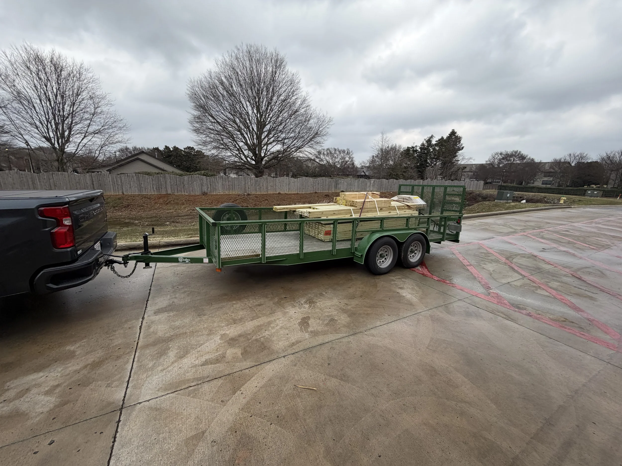 A black pickup truck pulling a green flatbed trailer with lumber on it in a parking lot, with trees and a cloudy sky in the background.
