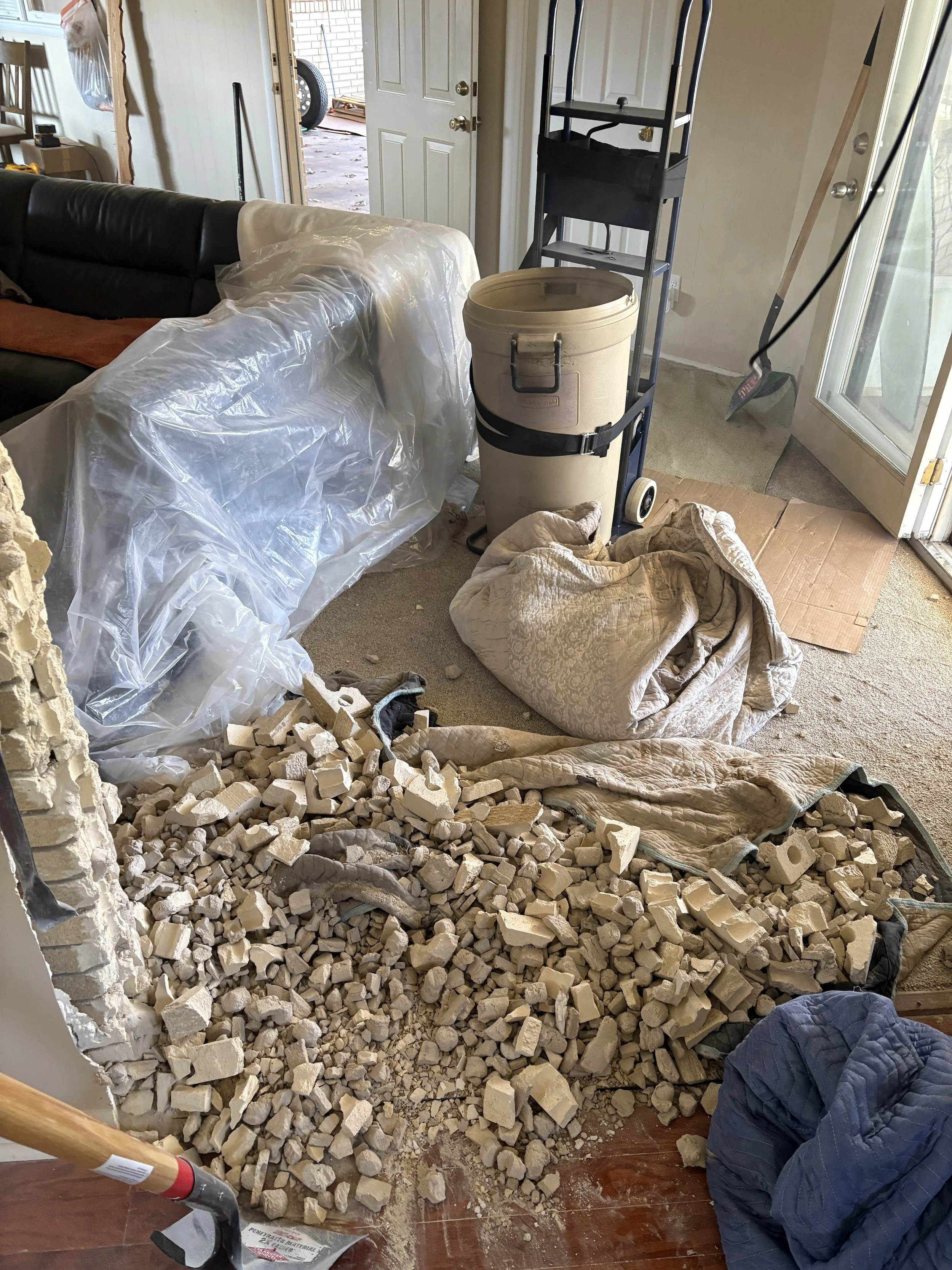 Debris and rubble on the floor of a room, with a partially demolished wall, a plastic cover over furniture, a bucket on a hand truck, a shovels leaning against a door, and various tools and items scattered around.