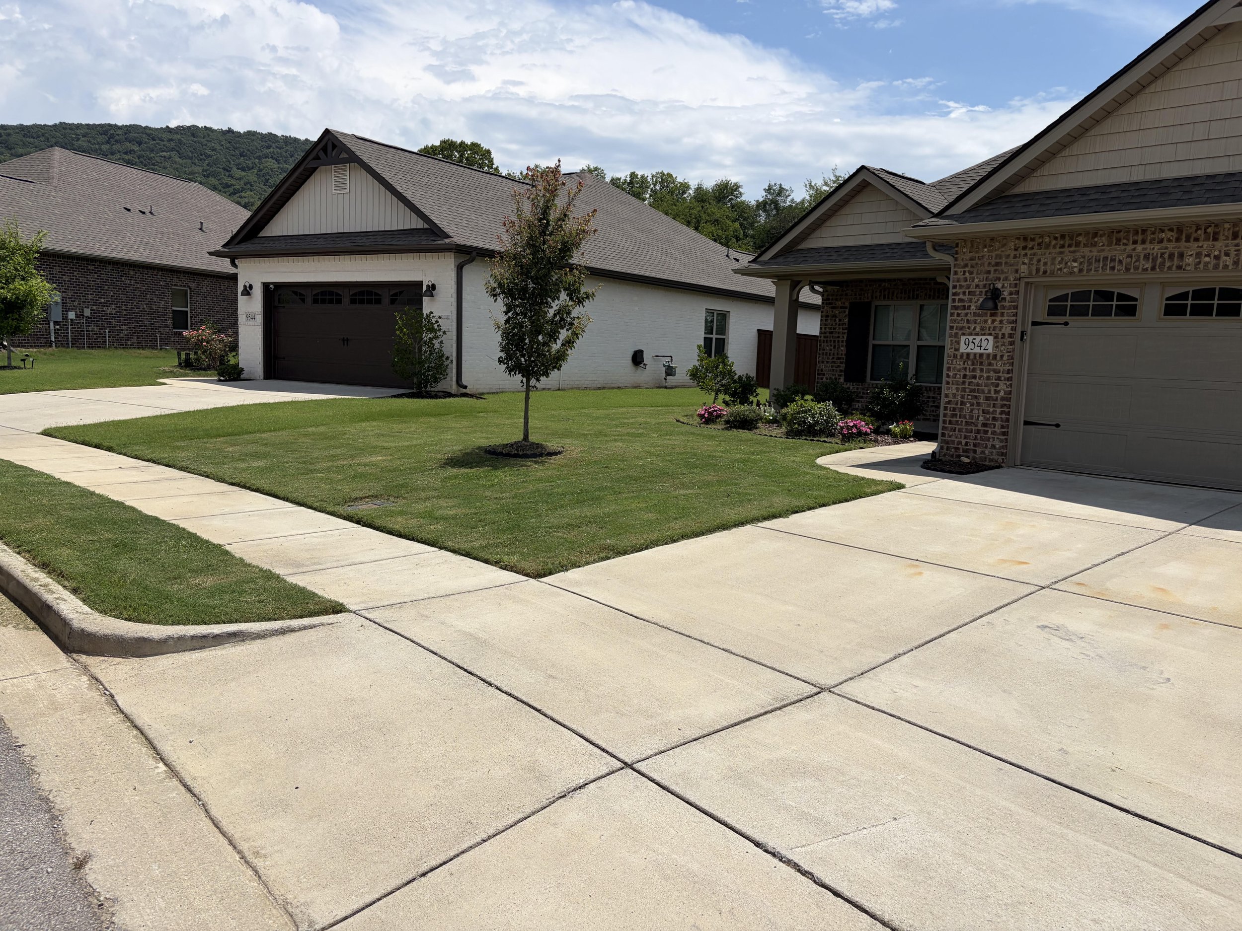 View of suburban houses with brick and white siding, pitched roofs, manicured lawns, small trees, and a clear sky with some clouds in the background.