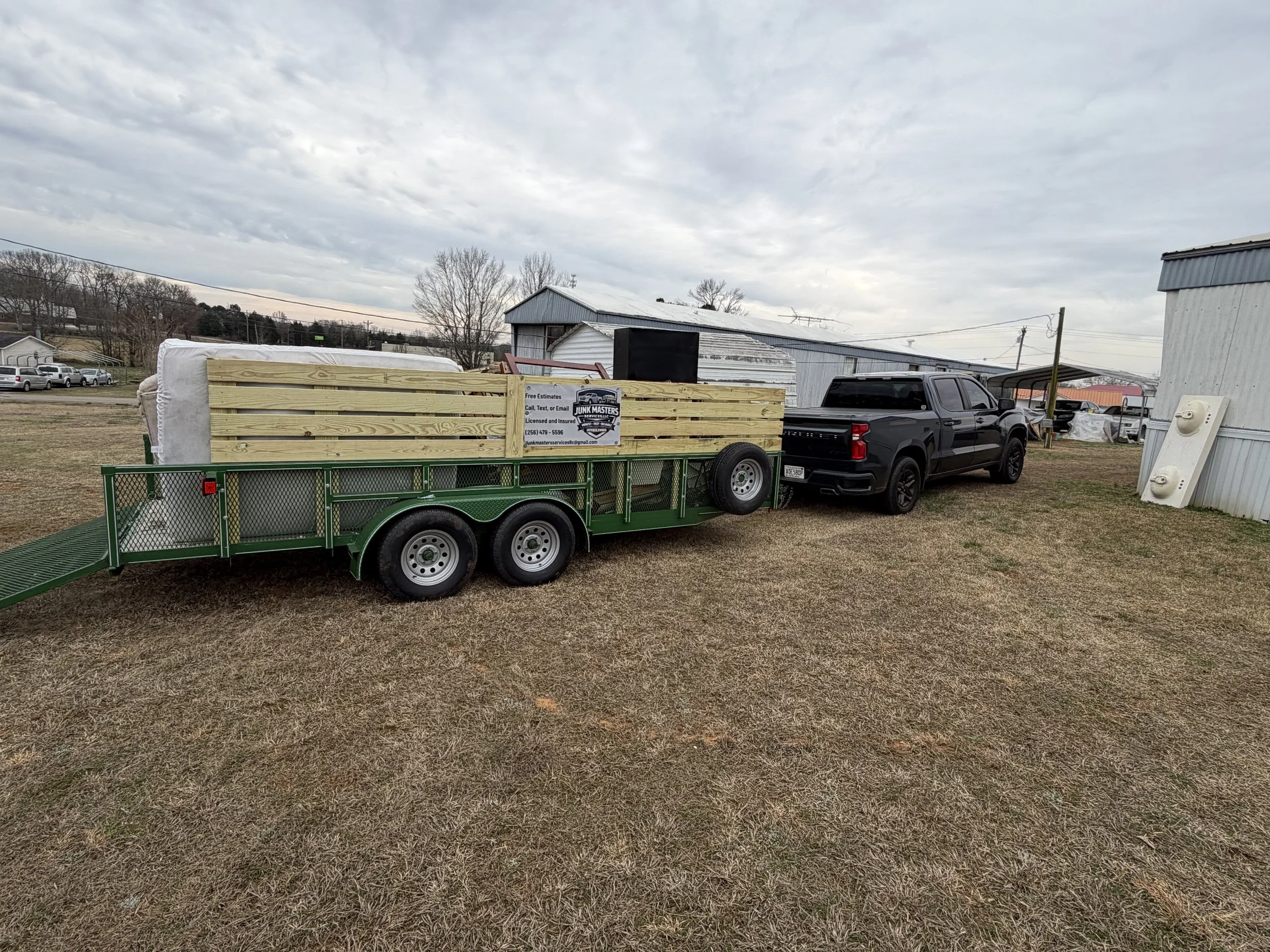 A black pickup truck pulling a green utility trailer loaded with wooden boards and other materials, parked on a grassy field with buildings and trees in the background under a cloudy sky.