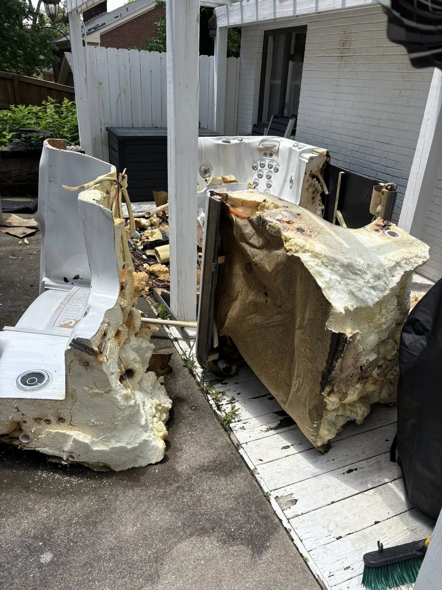 Disassembled hot tub or spa on a patio next to a white wooden fence and a house with a white brick exterior. Parts and insulation materials are scattered around.