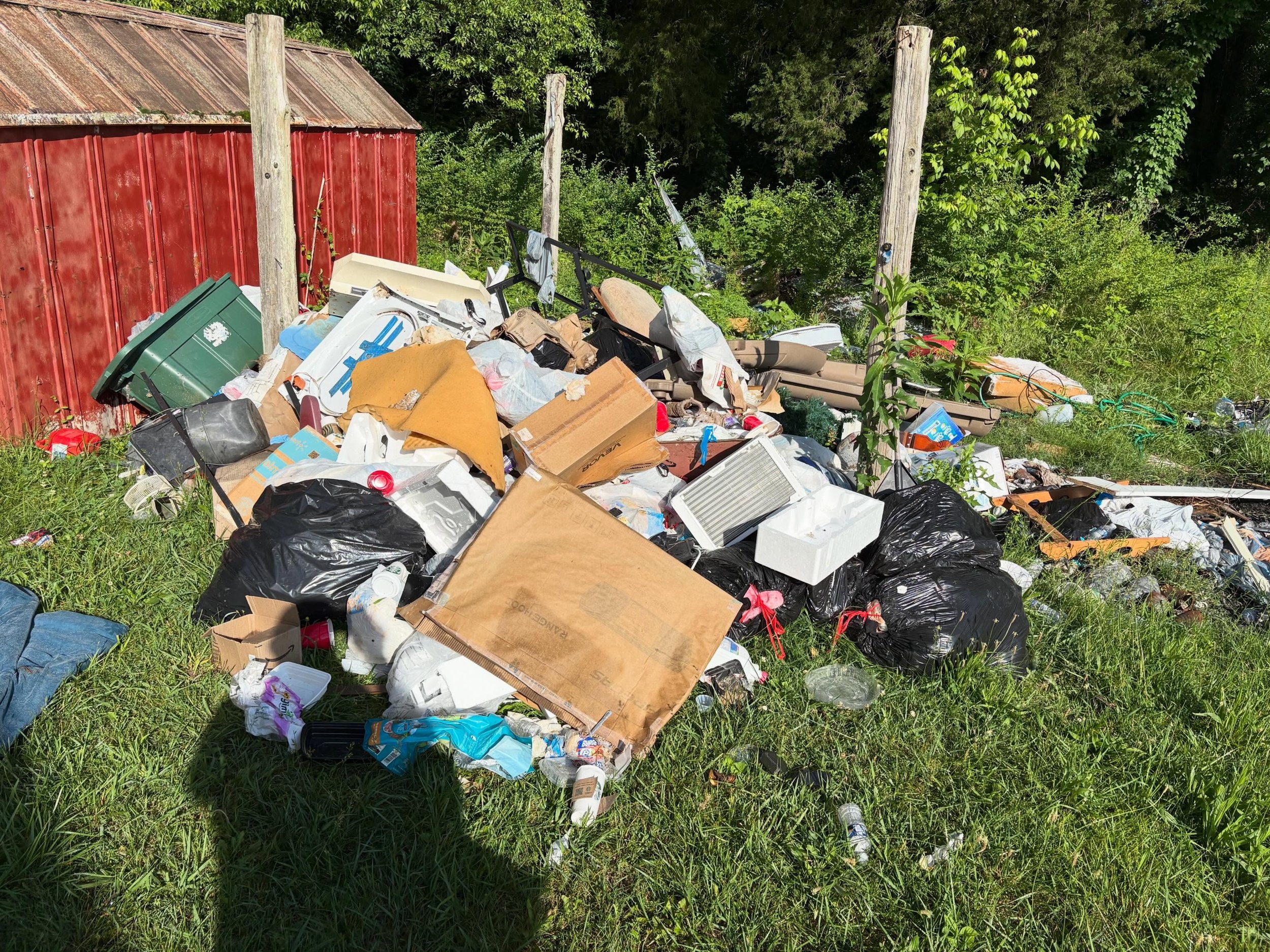 Pile of trash and debris scattered on grass next to a red shed and wooden fence with green trees in the background.