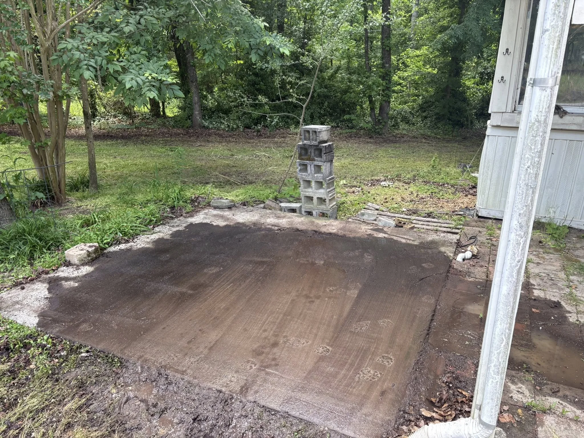 A backyard with trees and a grassy area, a small pile of cinder blocks, and a partially completed concrete patio or ground surface near a white house or shed with a leaning white door. There are some garden plants and patches of dirt.