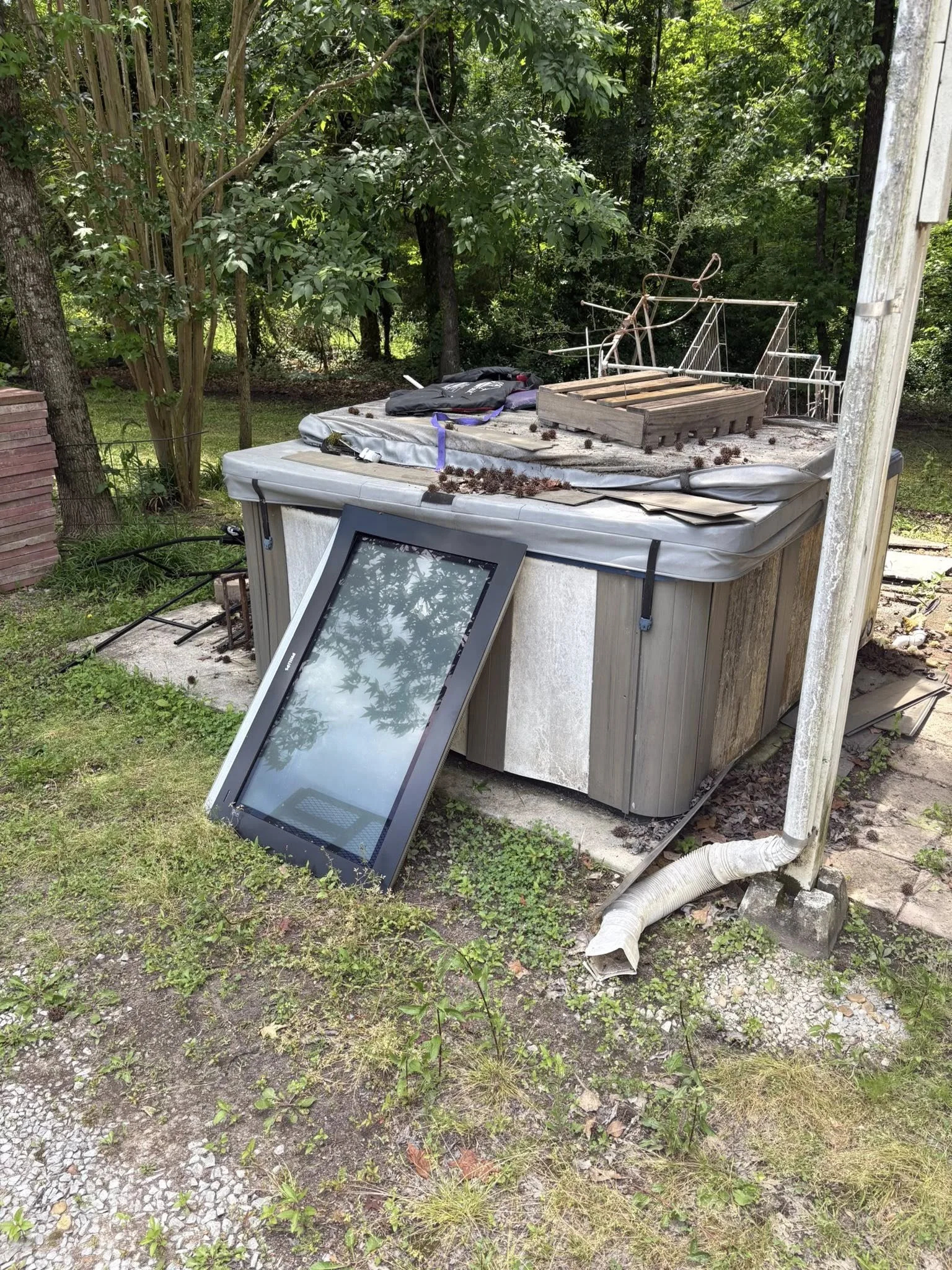 A backyard with a hot tub covered with a tarp, a broken window leaning against it, and an old, rusted bicycle beside a fenced wooded area.