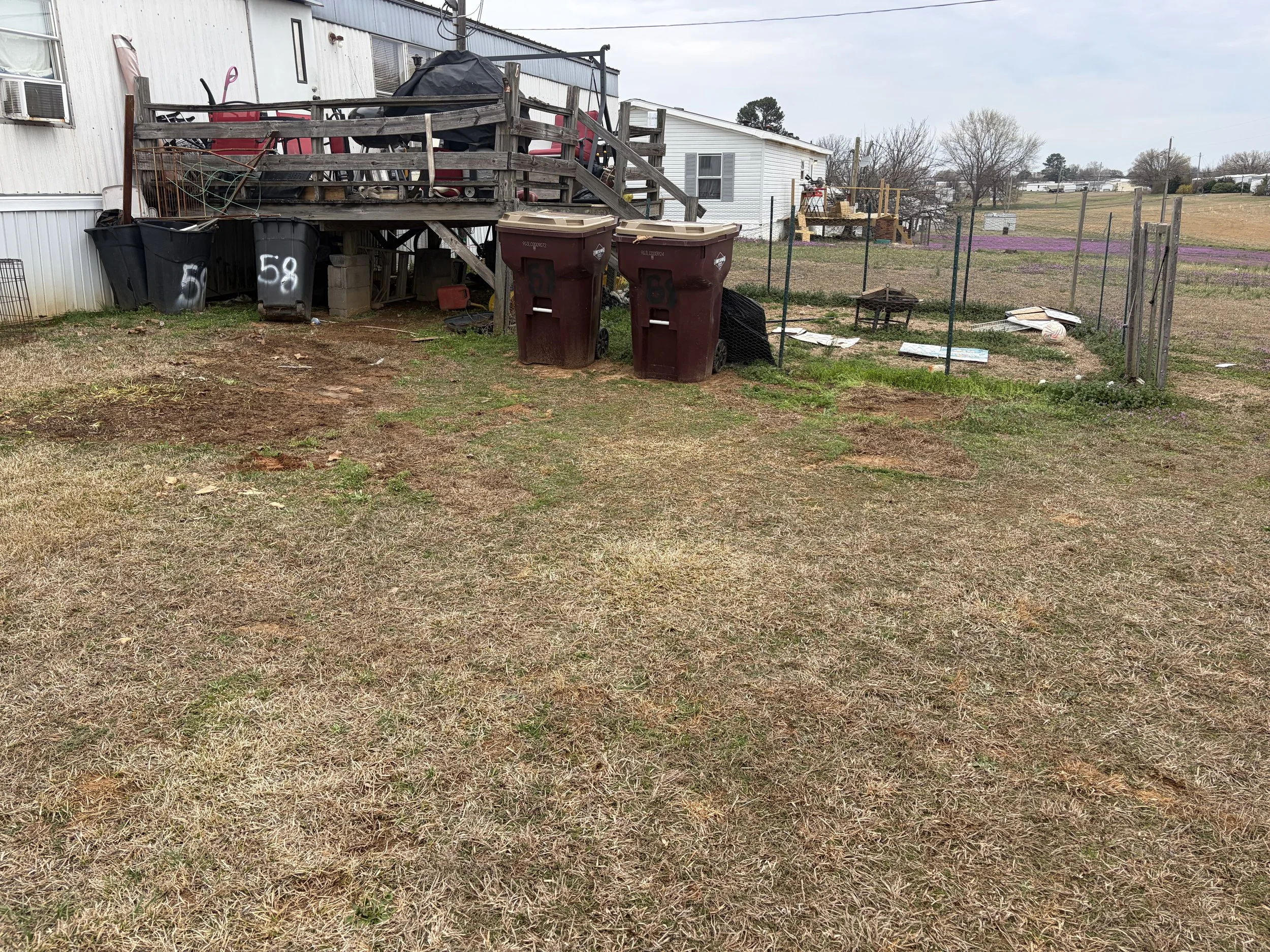 Backyard with a wooden deck, garbage bins, some debris, and a fenced area on a cloudy day.