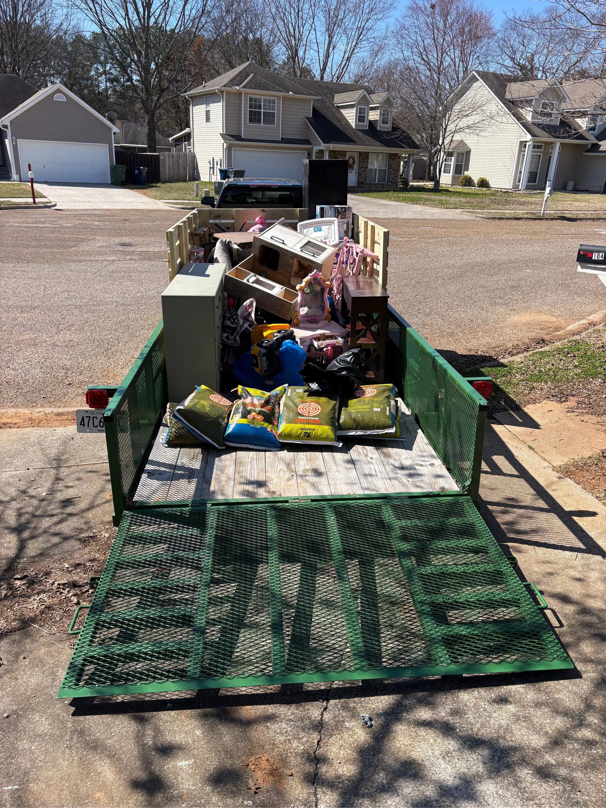 A green utility trailer filled with household items, including furniture, bags of yard waste, and assorted small objects, parked on a residential driveway under sunny weather.
