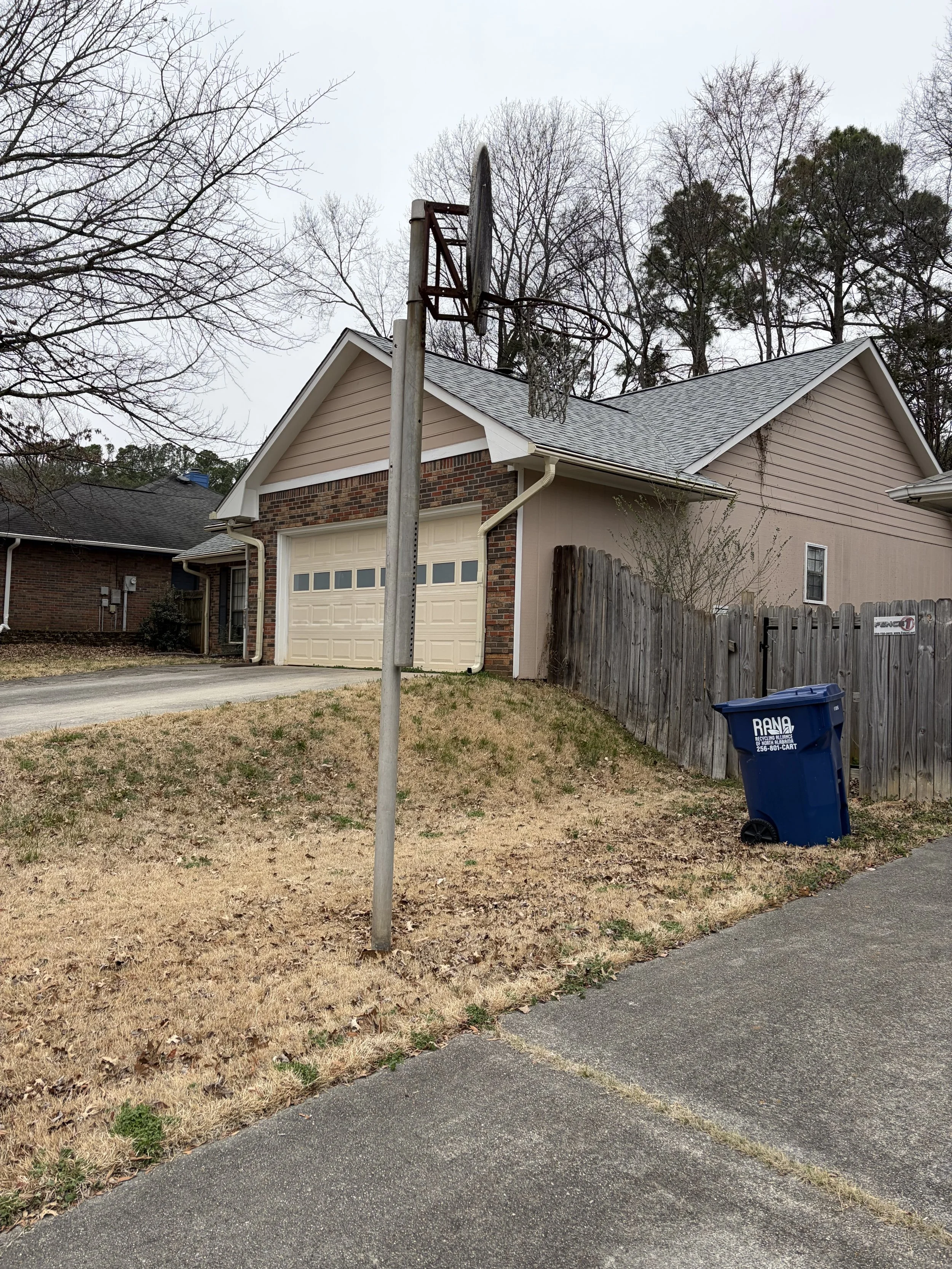A suburban house with a detached garage and a basketball hoop missing its backboard. The house has a brick and beige siding exterior, and there is a blue trash bin near a wooden fence. The yard has patches of grass and a concrete driveway.