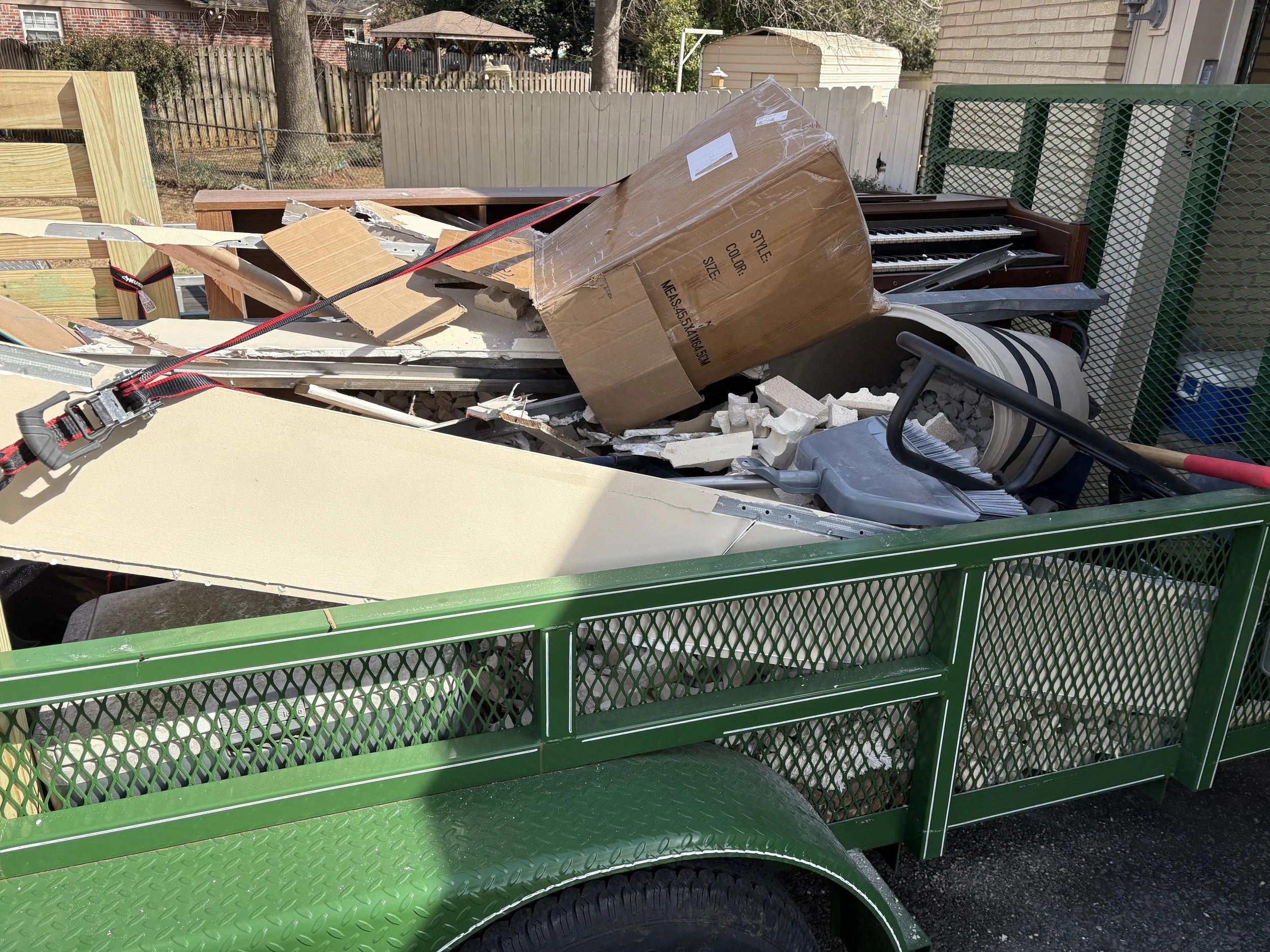 The image shows a green utility trailer filled with debris including a cardboard box, wooden planks, foam, a plastic chair, and part of a piano, parked outdoors with a backyard and fence in the background.