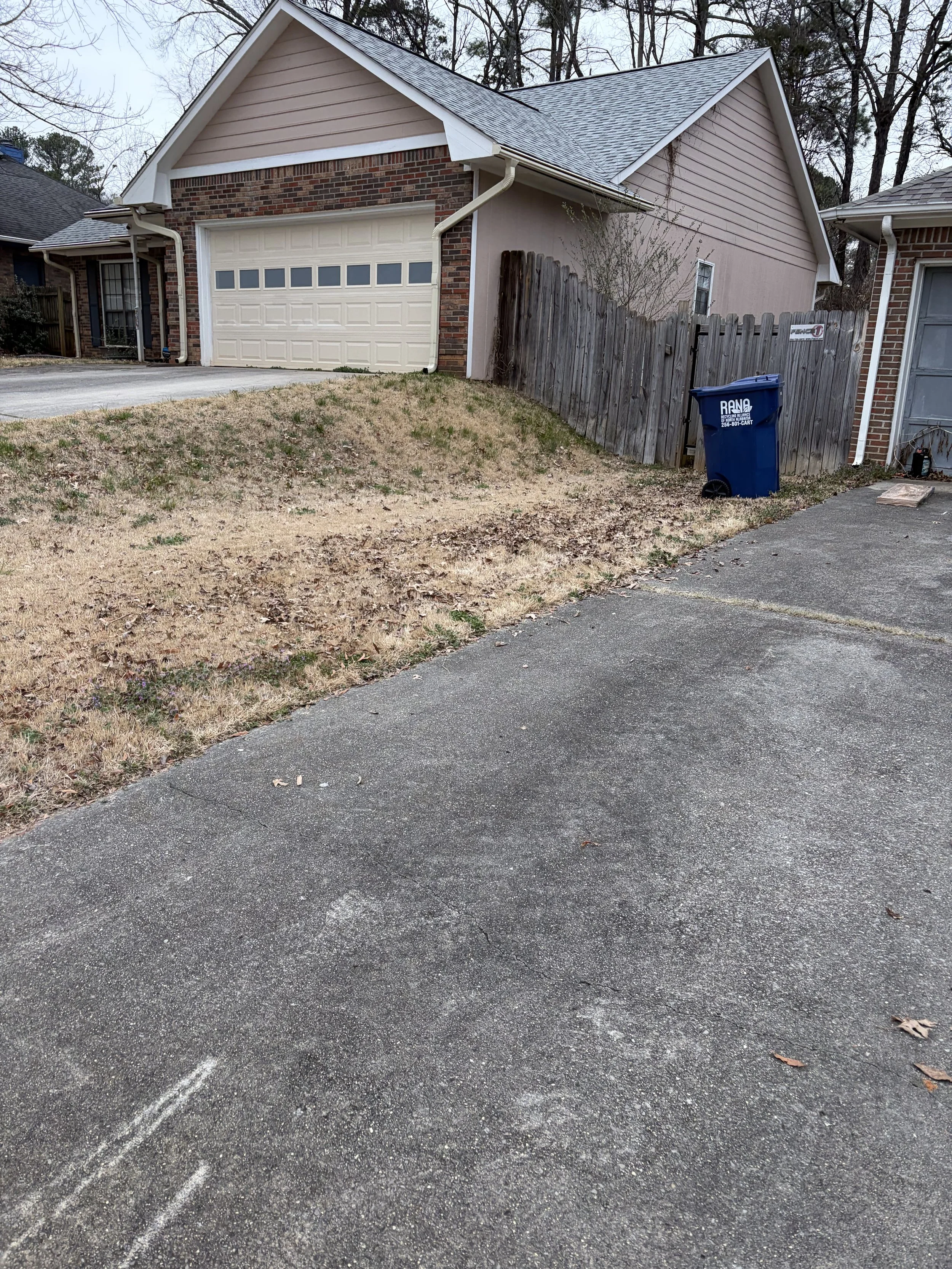 A suburban house with a garage, a small lawn area, and a wooden fence. A blue trash bin is near the fence, on the driveway.