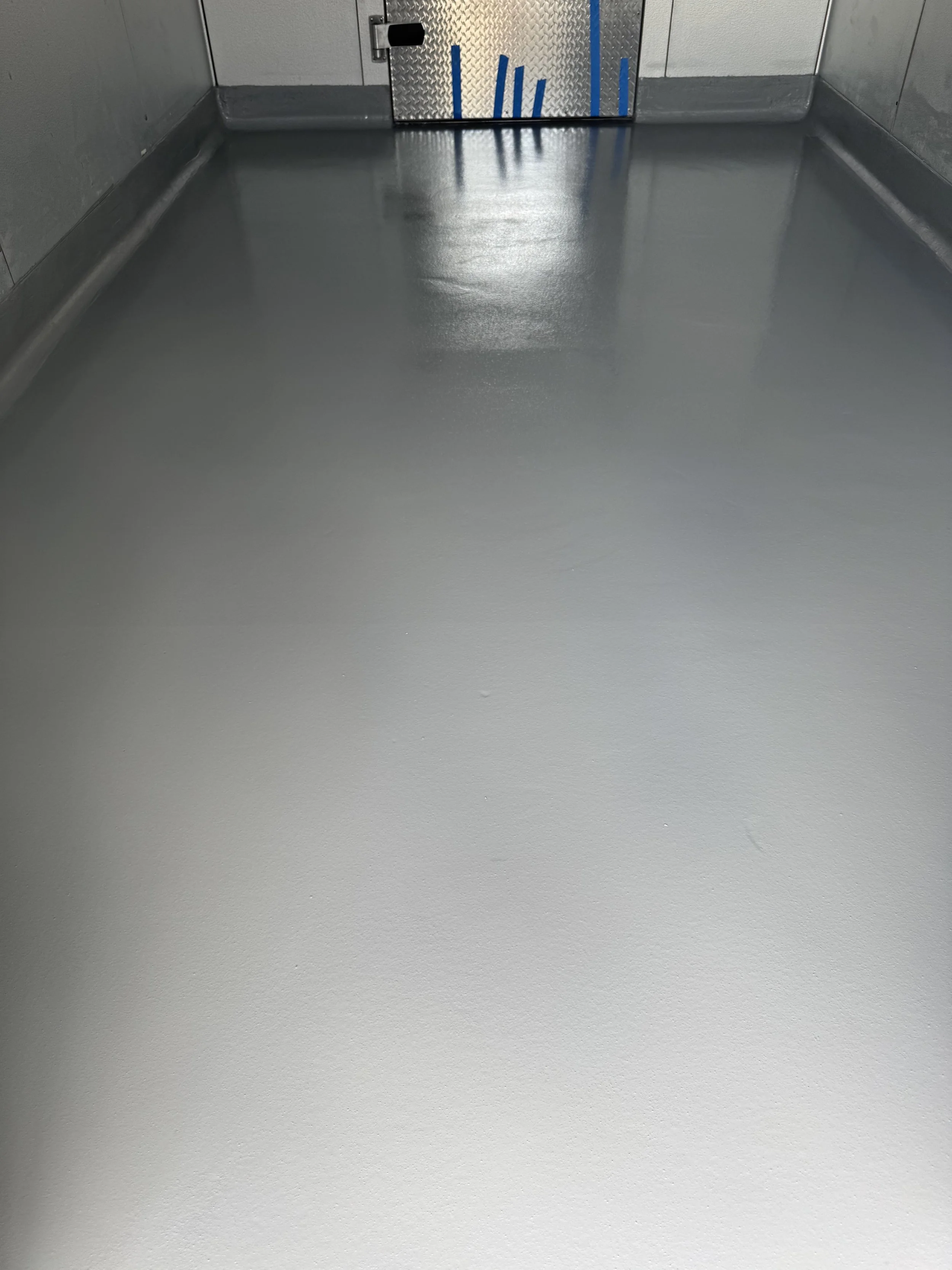 A freshly painted, solid gray concrete floor inside freezer in a restaurant, with a metal door at the far end. epoxy, polyaspartic, polyurea