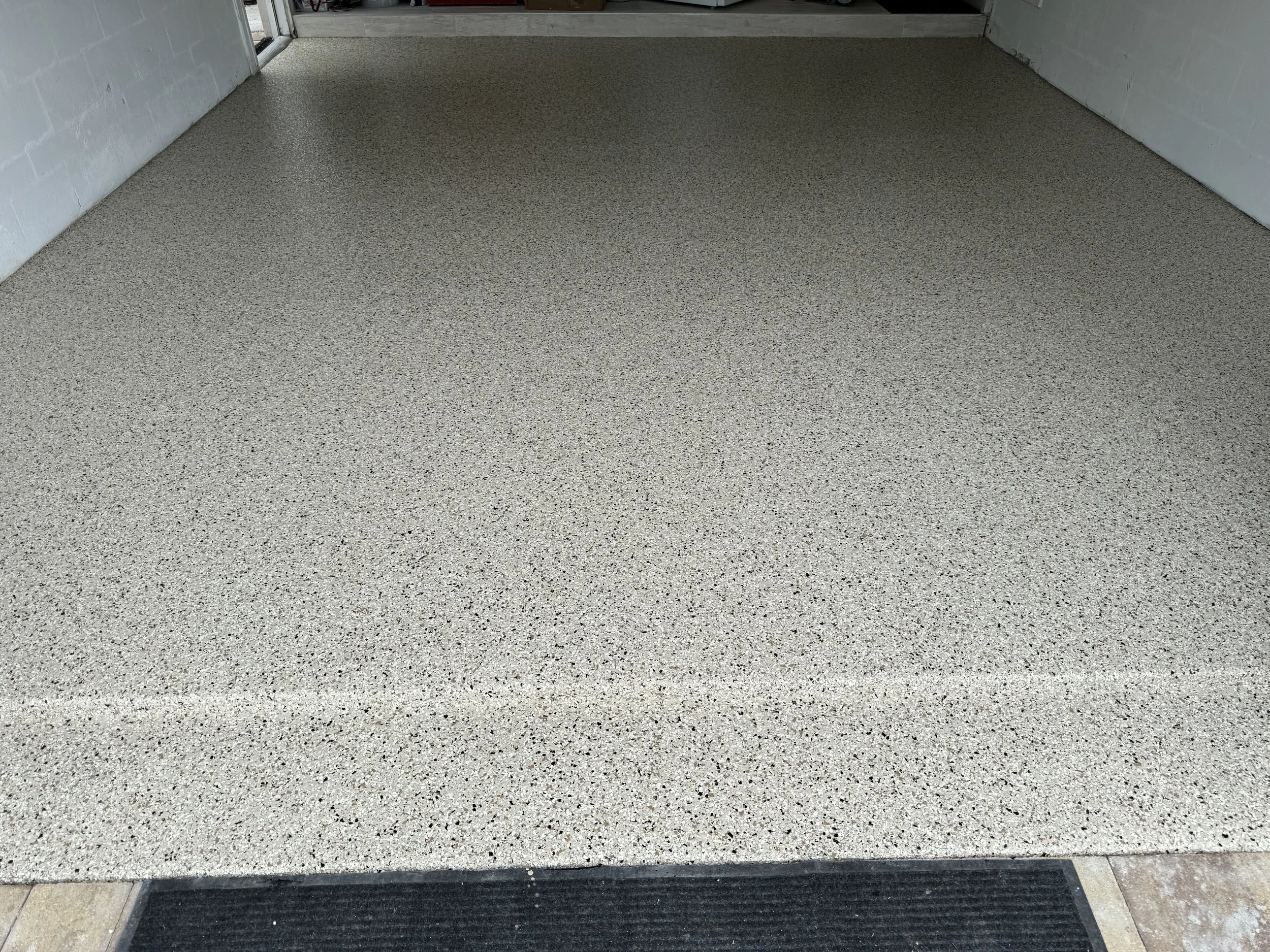 A freshly installed beige speckled epoxy coated garage floor with a black rubber mat at the entrance. polyaspartic garage floor, polyurea