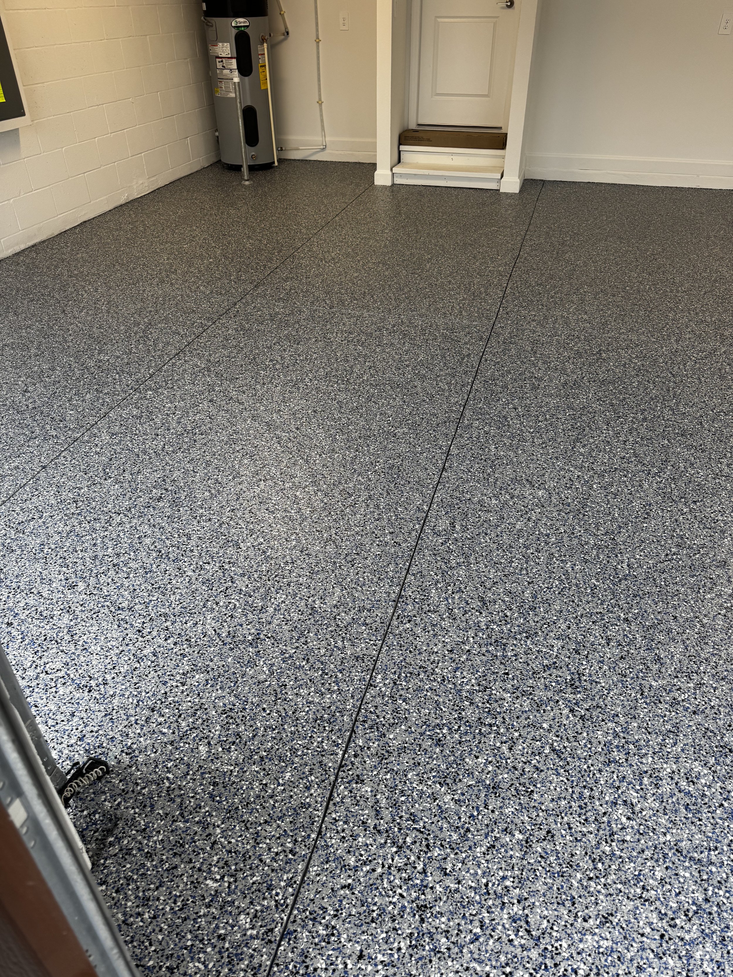Interior of a garage with speckled grey and black epoxy flooring, a white brick wall, a water heater, and a closed door.
