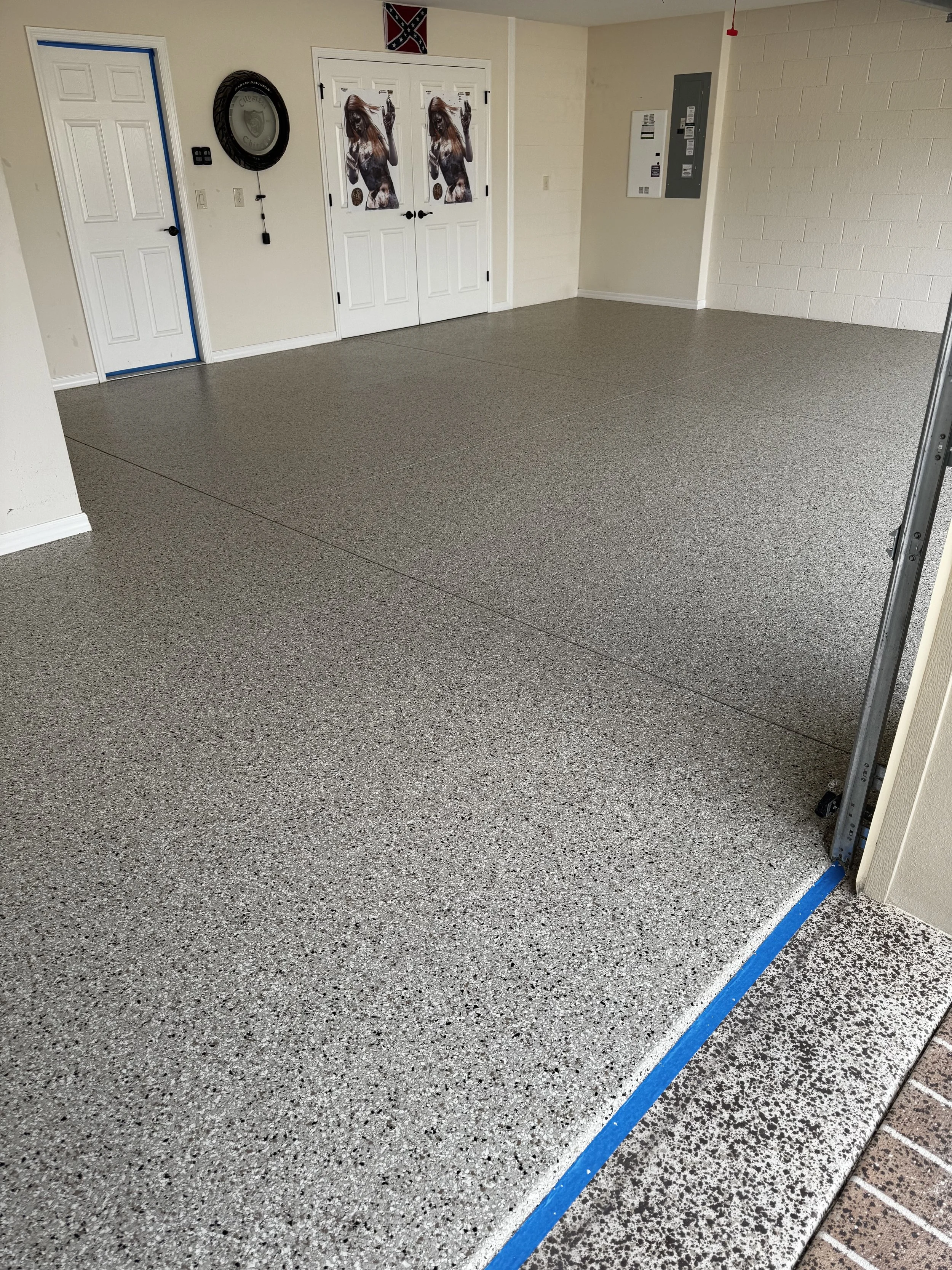 Interior view of a clean garage with a speckled epoxy floor, white walls, and a white door with a horse-themed curtain. There is a circular wall clock, a Confederate flag, and electrical panel on the walls. epoxy, polyaspartic, polyurea, tampa bay