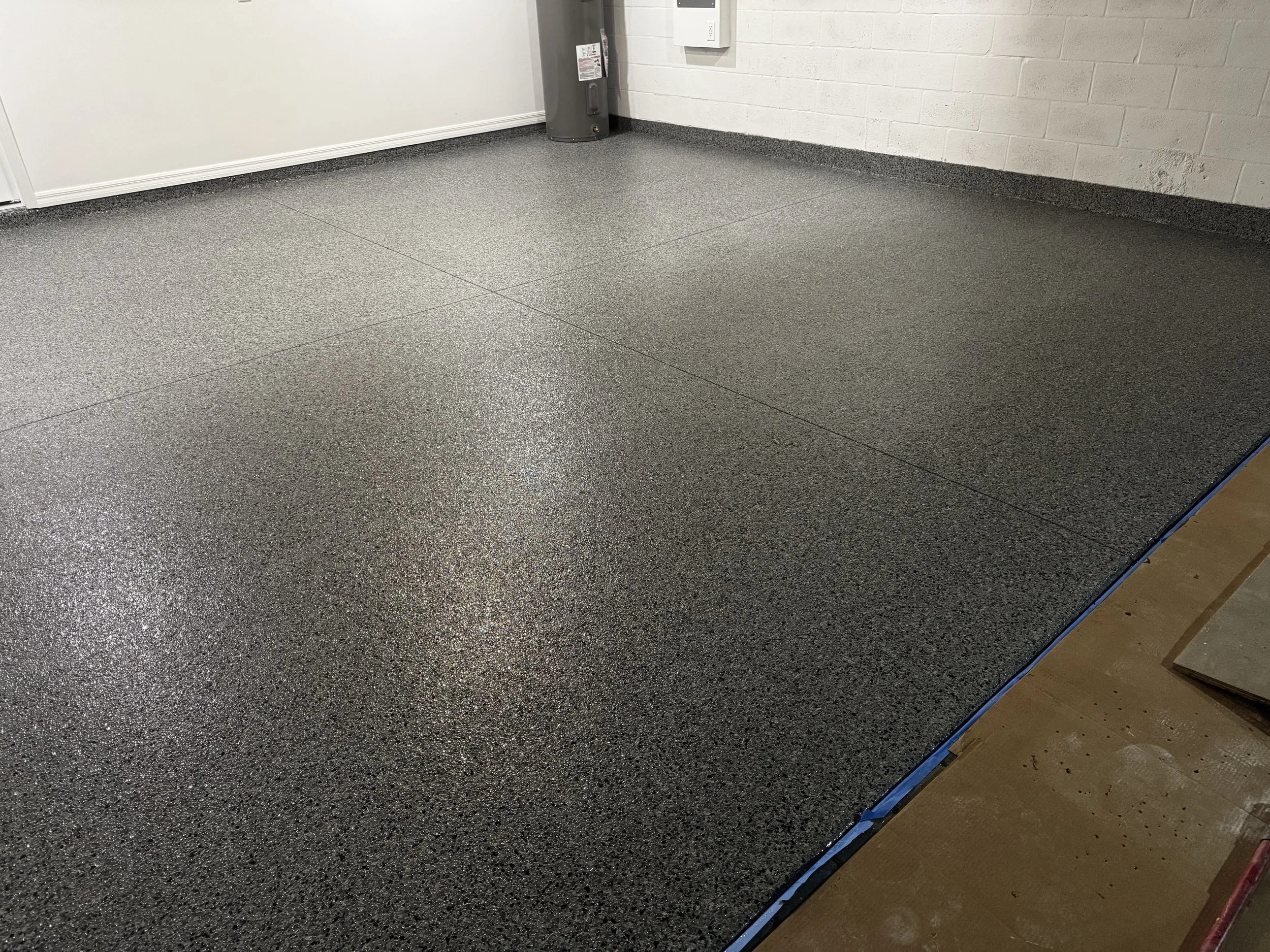 Newly installed speckled gray epoxy flooring in a garage, with a water heater in the corner and a wooden floor partially visible at the side. polyaspartic, polyurea