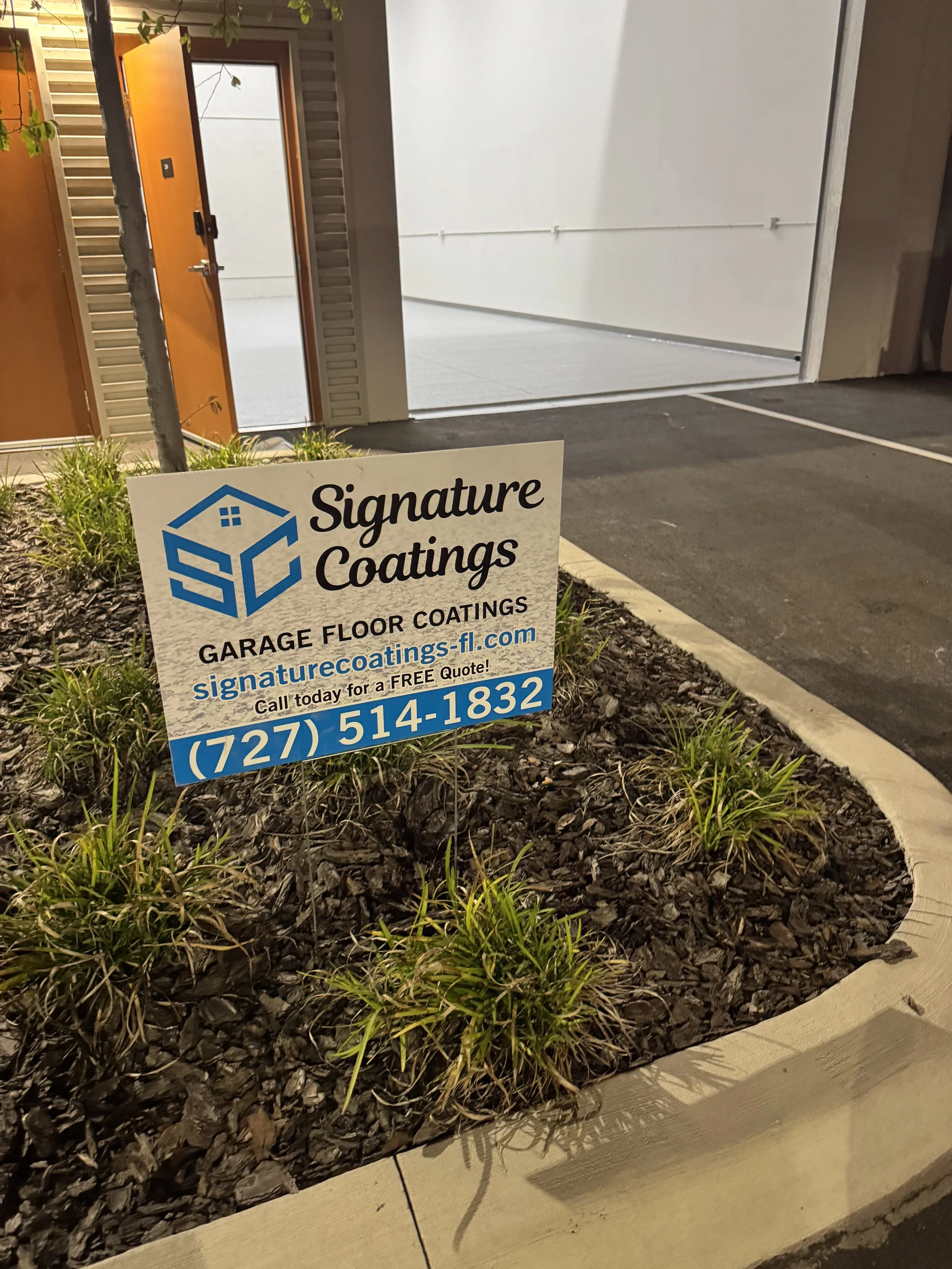 Sign for Signature Coatings advertising garage floor coatings, with a phone number and website, placed in a landscaped area outside a building with an open garage door.