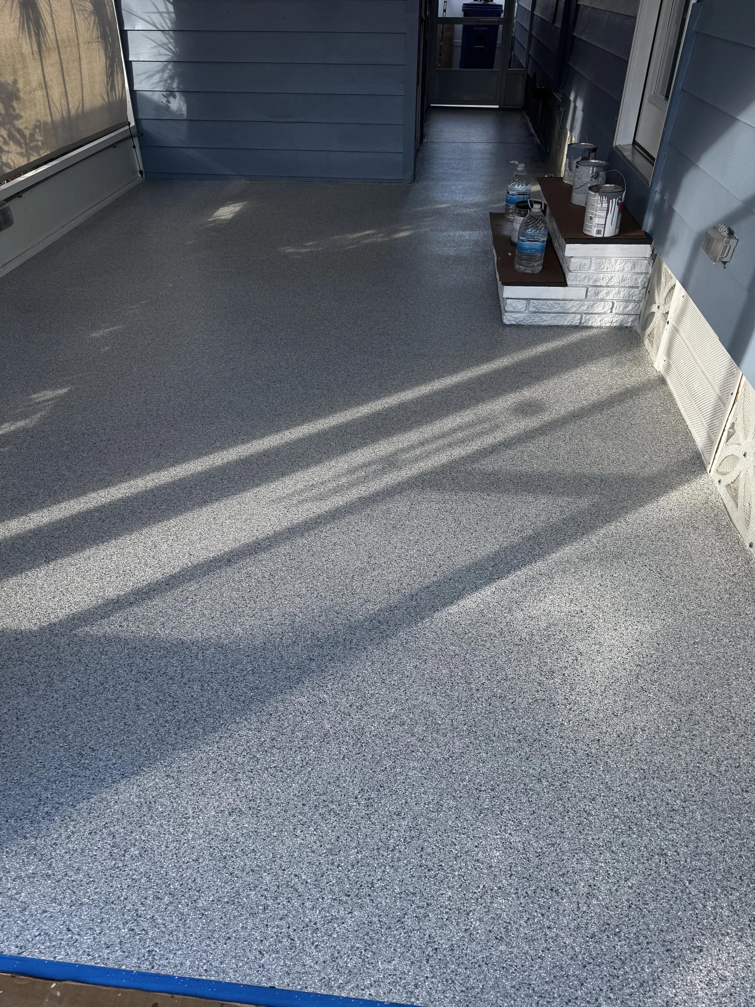 Residential porch with newly coated, textured flooring in gray, blue siding, and various bottles and cans placed on a small ledge near the wall. epoxy, polyaspartic, polyurea
