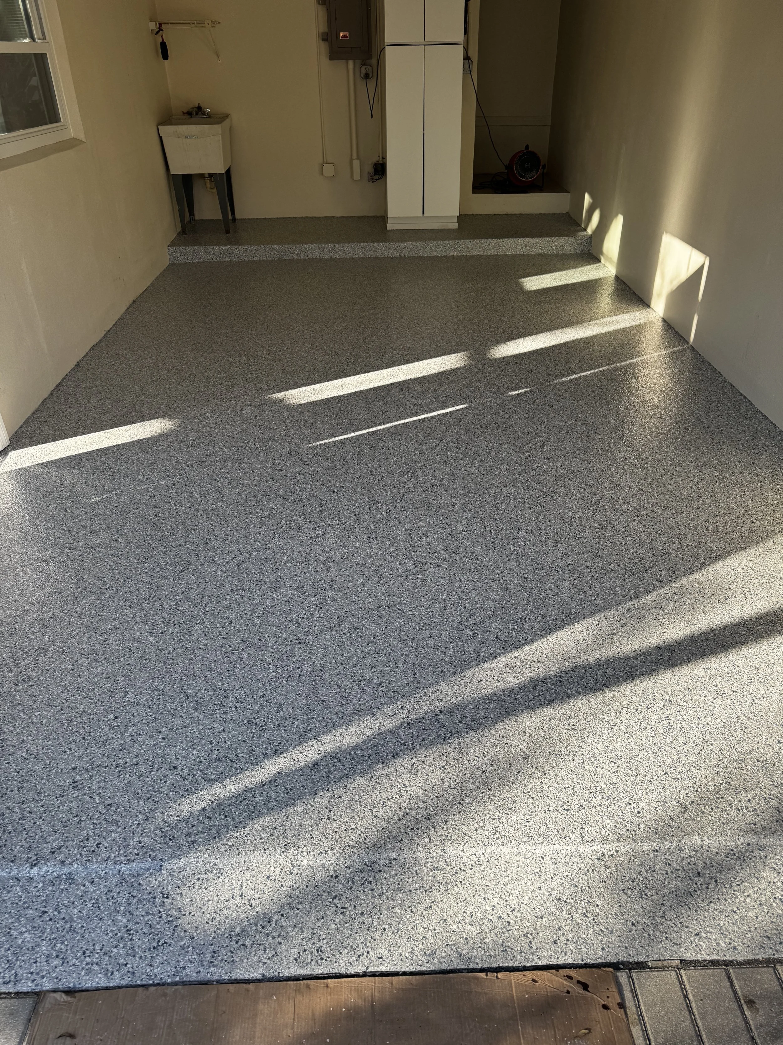Residential garage with speckled gray epoxy flooring, sunlight casting shadows on the floor, small utility sink on the left, some electrical panels and wires on the back wall, and a portable heater in the back right corner.
