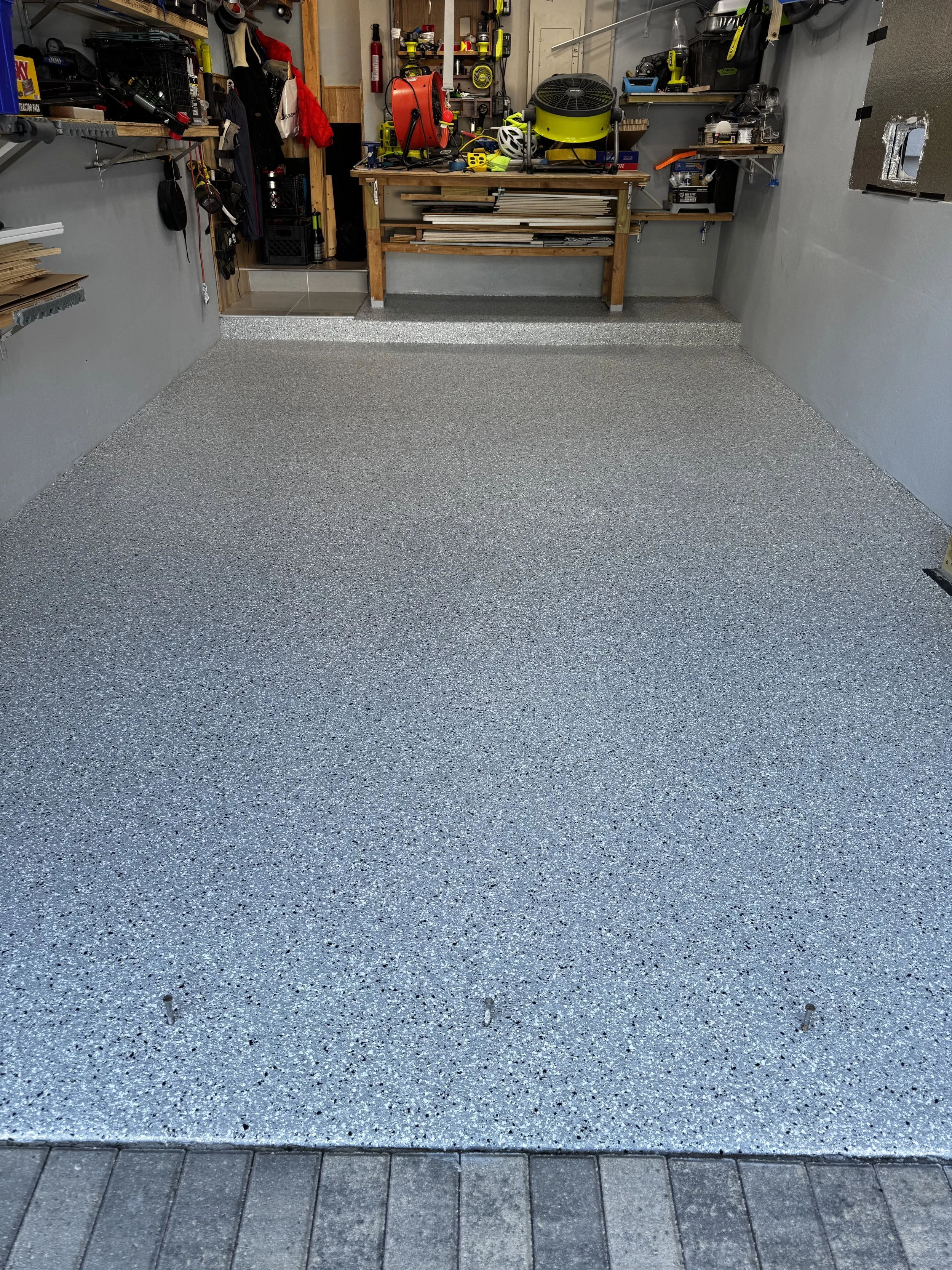 A clean and organized garage with a speckled epoxy floor, tools and equipment on shelves, a workbench with various items, and a doorway leading to another room.