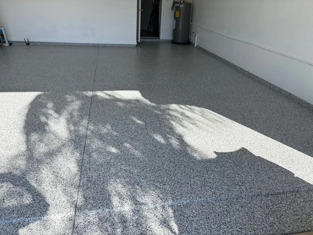 Empty garage with speckled epoxy floor, a water heater, and shadows of a tree cast on the floor.