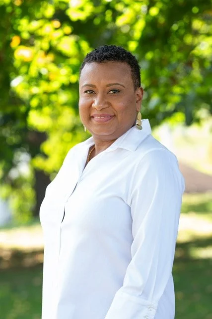 Ria Savoy, birth and postpartum doula in Philadelphia County wearing a white shirt standing outdoors with green trees in the background.