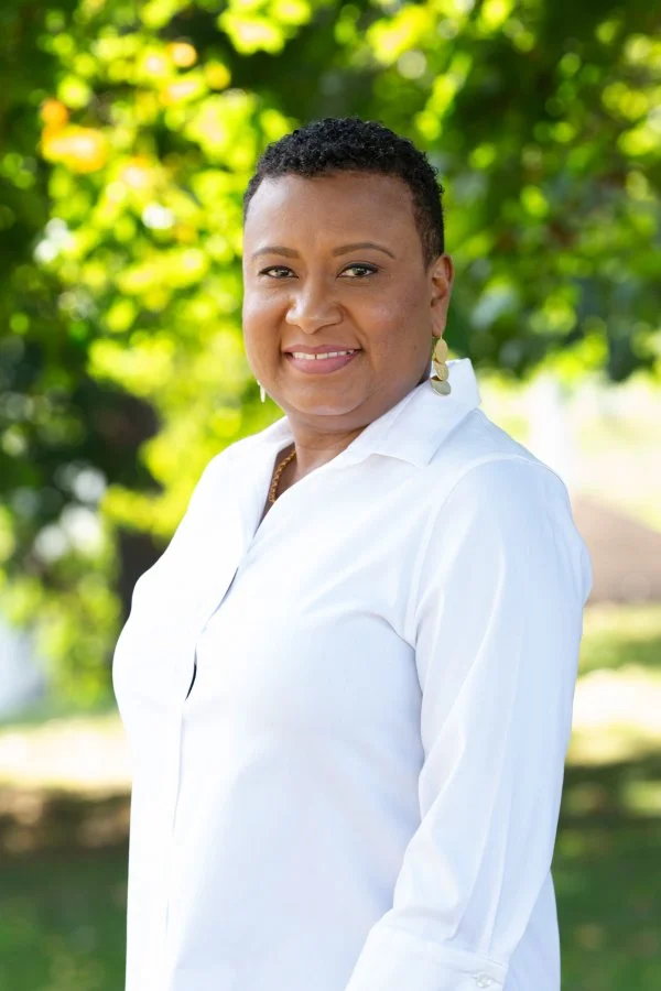Ria Savoy, postpartum doula and sleep consultant, in a white shirt standing outdoors with green trees in Montgomery County, PA.